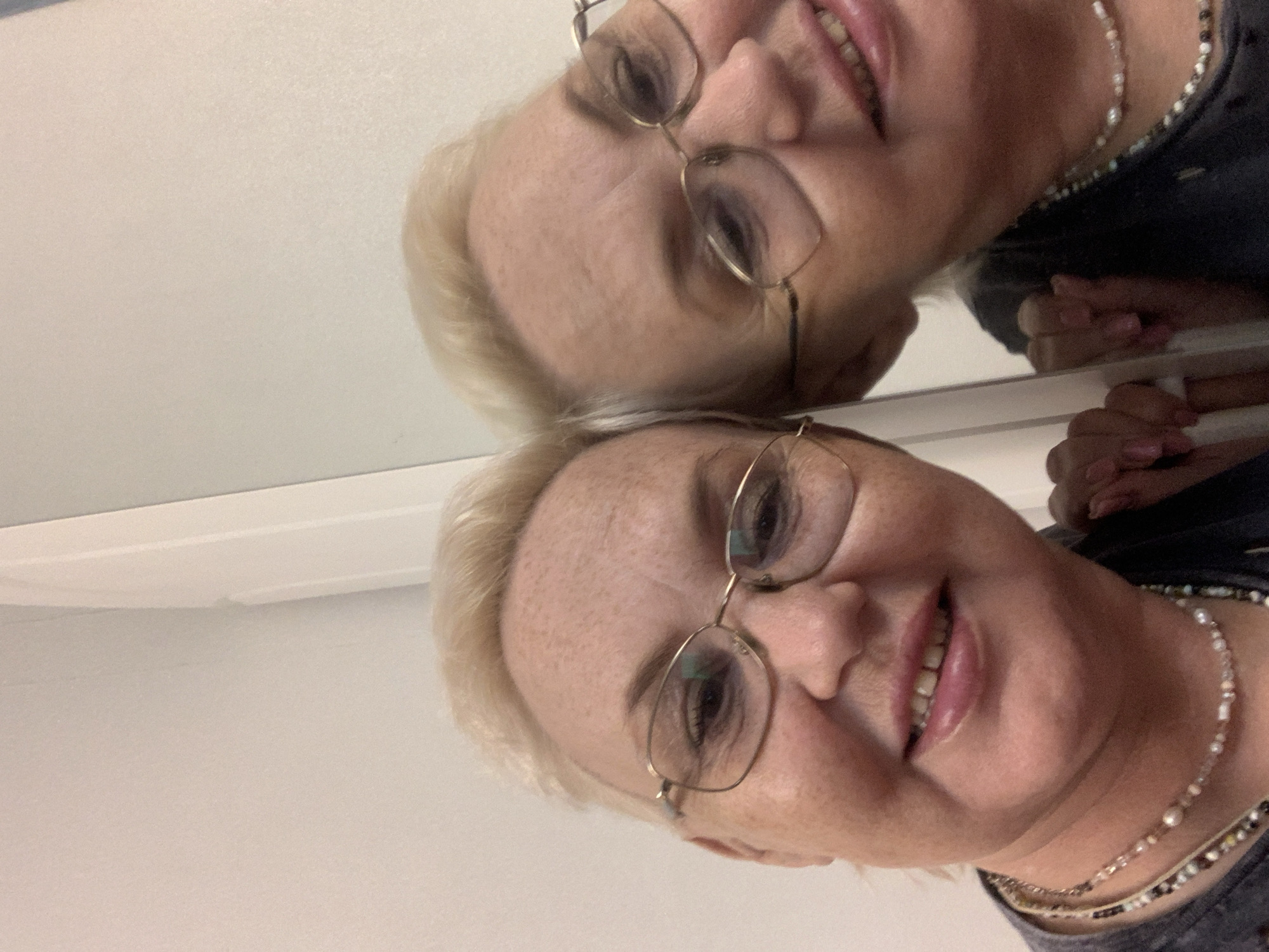 A woman with fair skin and blonde hair wearing glasses and a beaded necklace poses for a selfie in front of a mirror. Her reflection is visible in the mirror above her. The background appears to be a plain, light-colored wall. The location, Chaikovsky, Russia, cannot be verified from the provided image.