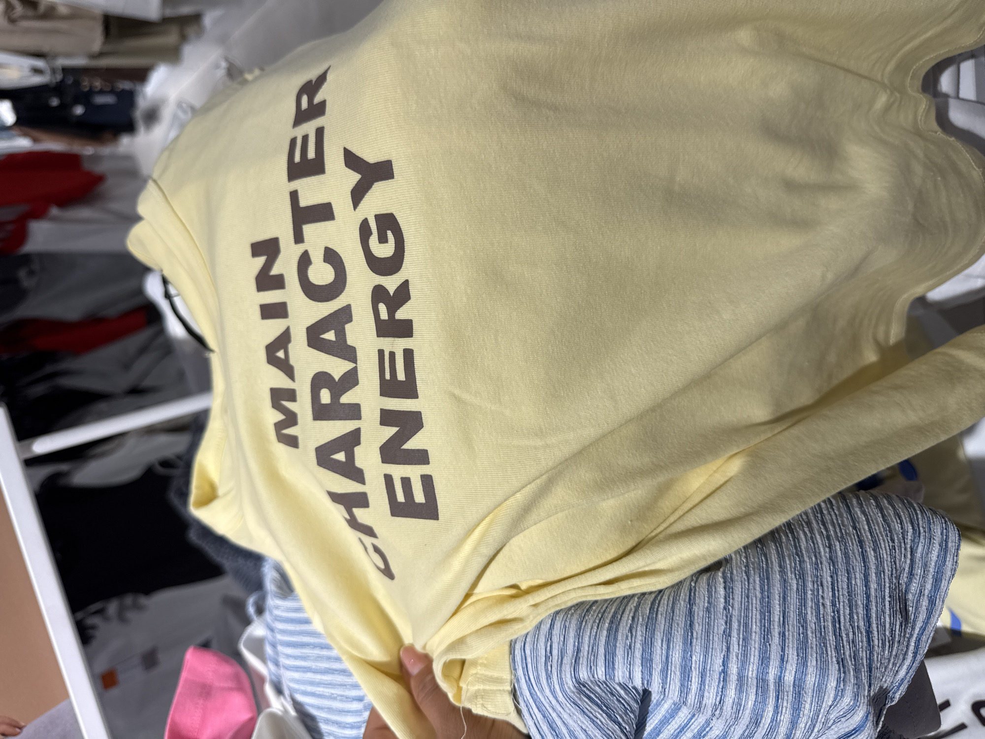 The image shows a close-up of a light yellow t-shirt with dark brown text that reads "MAIN CHARACTER ENERGY". The t-shirt is draped over other folded clothes, including a blue and white striped item. A hand is visible holding the t-shirt from underneath. The background is blurred but appears to be a retail setting, possibly a clothing store or a closet, with shelves and other garments visible. The lighting suggests it's indoors. No specific time of day, weather, or emotional context can be determined from the image.