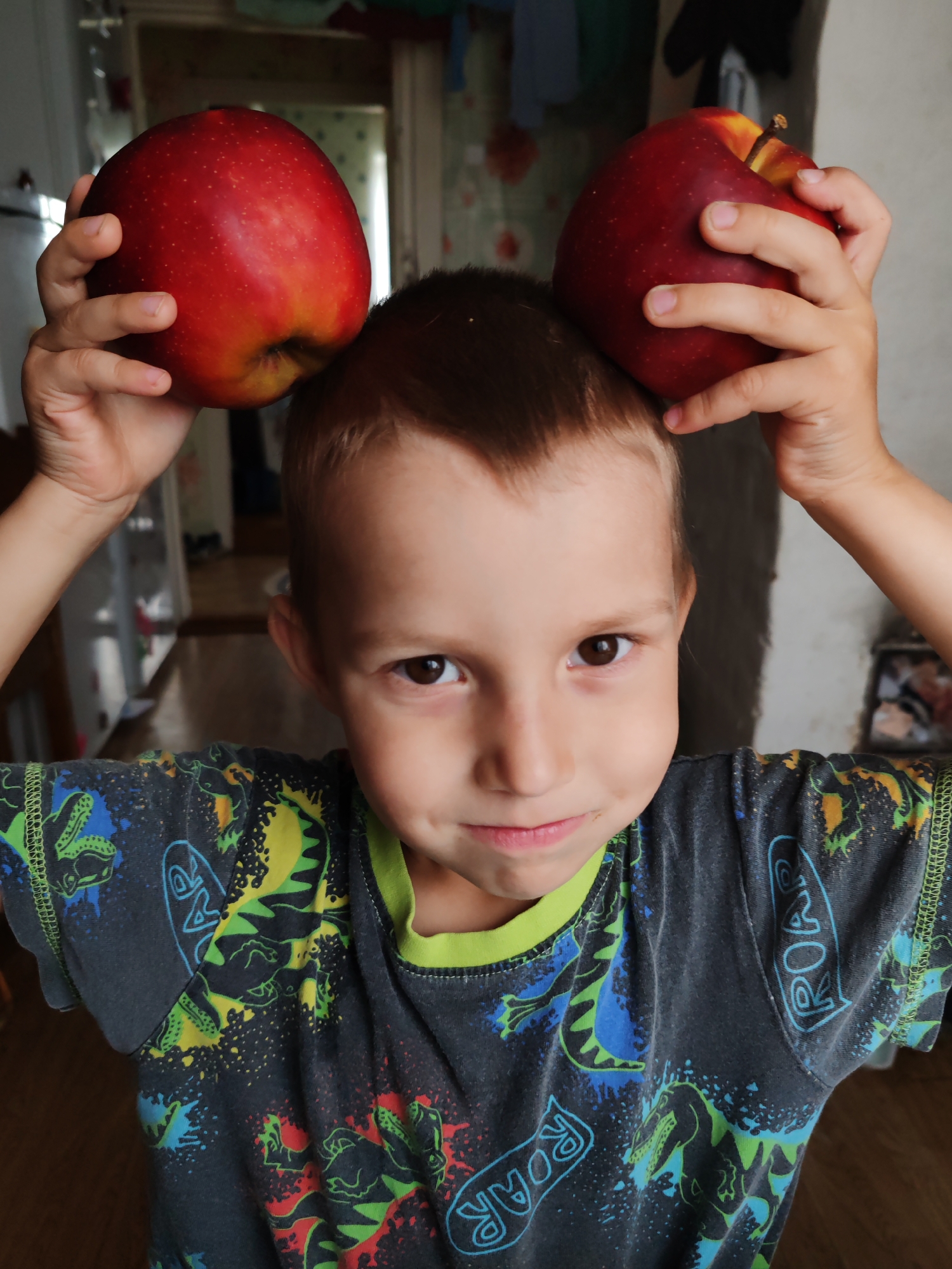 A young boy with brown hair and brown eyes is holding two red apples, one on top of his head with his left hand, and another with his right hand. He is wearing a dark grey t-shirt with a green collar and a repeating dinosaur pattern. The background appears to be an indoor setting, possibly a home, with wooden furniture and what looks like a doorway or hallway visible. The lighting suggests it is daytime. The image was taken in the city of Chuhloma, Russia.