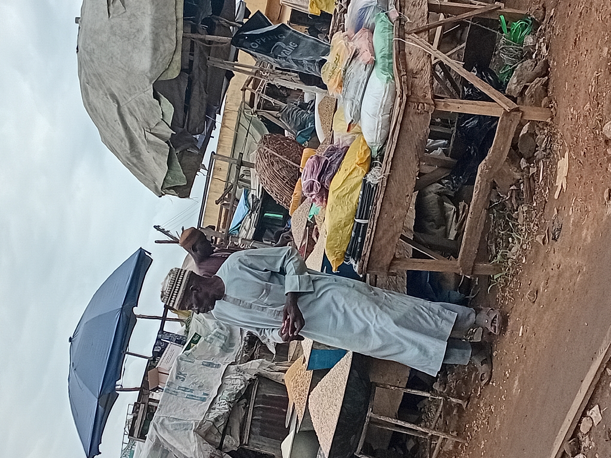 The image depicts an outdoor market scene in Maiburiji, Nigeria. Two men are present. The primary subject, on the left, is wearing a light blue kaftan and a patterned cap. He stands facing the right, holding what appears to be a small item in his hands. To his right and slightly behind him, another man, wearing a reddish-brown shirt and a cap, is partially visible, looking downwards.
The foreground is dominated by a wooden stall laden with various goods. Piles of grain or seeds are displayed in large metal bowls. On the stall's surface, there are stacks of bagged goods in different colors, including yellow, white, green, and purple. A large, worn, beige tarp covers an overhead structure, providing shade. To the left, a dark blue umbrella is propped up, also offering shade. The ground is covered in dirt and small stones, with some sparse vegetation. In the background, more market stalls and structures are visible, along with sacks of goods and what looks like a hanging black bag with white lettering. The sky is overcast.