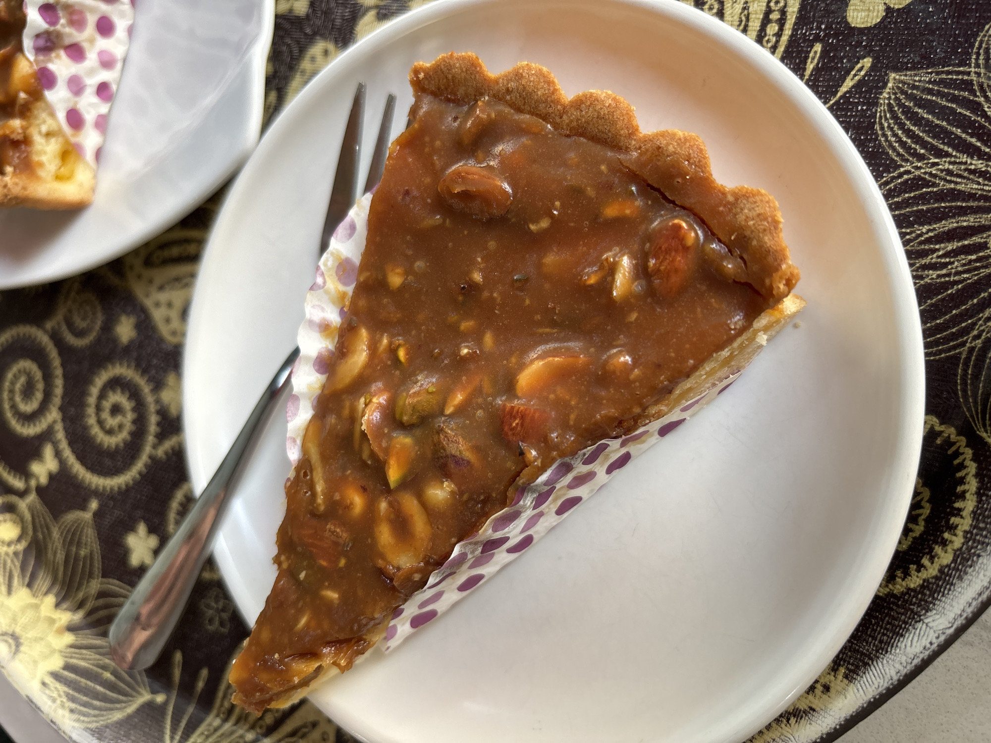 A triangle slice of nut tart is on a white plate, with a fork alongside. The tart has a golden-brown crust, and a caramel-colored nut filling containing almonds and pistachios. A cupcake liner with purple polka dots surrounds the base of the tart. The plate sits on a patterned placemat in shades of brown and gold. Another plate with a slice of tart is partially visible in the background. The location cannot be determined.
