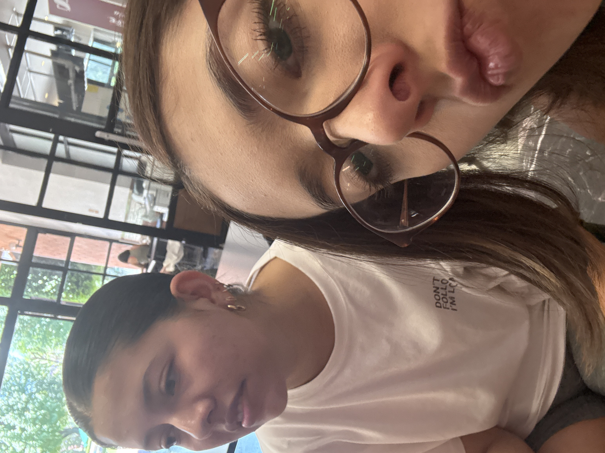 Two young women are in a well-lit indoor space, likely a restaurant or cafe, given the window view suggesting an outdoor area with trees. The woman closer to the camera is wearing glasses and making a duck face, while the other woman is looking downward. The woman in the foreground wears a white shirt with lettering that says "DON'T FOLLOW, I'M LOST". The location can be inferred by the context of the city mentioned in the request. No further details of San Andrés Cholula, Mexico, are visible in the image.