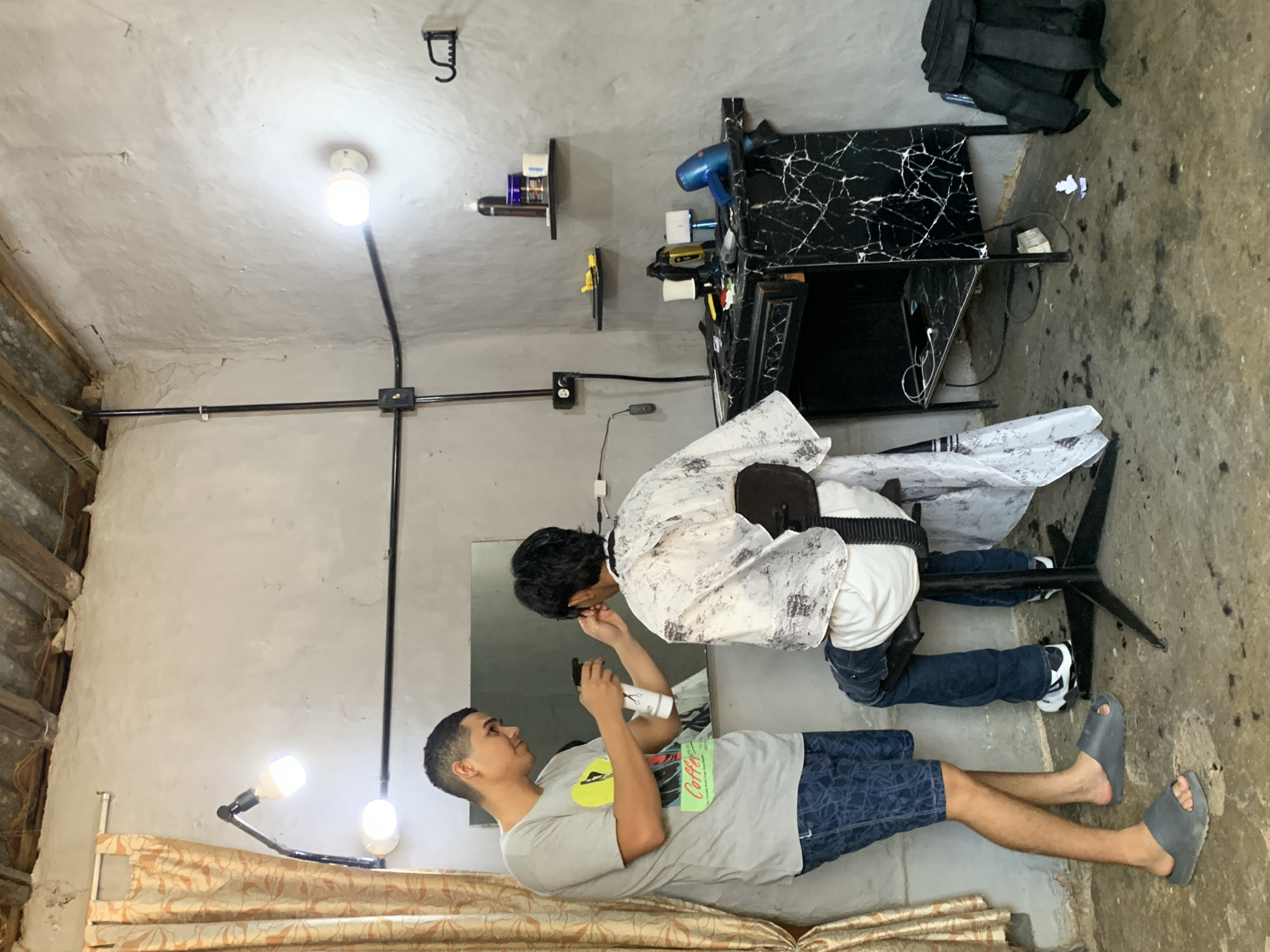 This image captures an indoor scene at a barbershop or grooming station in La Tagua, Venezuela. Two males are present: a barber and a client. The client, a man wearing a grey t-shirt with a yellow graphic and a green label displaying the word "Coffee," blue patterned shorts, and grey slides, stands while looking towards the barber. The barber, seated and wearing a white patterned smock over a white shirt and dark pants with black and white sneakers, is actively grooming the client's facial area or hairline, holding a white spray bottle and a small tool. Both individuals appear calm and focused on the grooming process.
The setting is functional and modest. The walls are light grey, textured, and appear to be unpainted concrete or plaster, while the floor is rough concrete. Electrical conduit pipes run along the ceiling and walls, with visible light bulbs providing illumination; one is on the ceiling, and two are on an adjustable lamp structure near the client. A rectangular mirror is positioned behind the client and barber.
To the right, a black desk with a white marble-like pattern serves as a barber station, holding a blue hairdryer, a yellow tool, and other grooming products. Below the desk, a black backpack rests on the floor. Two small black shelves on the wall display various bottles and containers. Patterned beige and brown curtains are visible on the left side of the frame, contributing to the practical, local establishment's ambiance.
