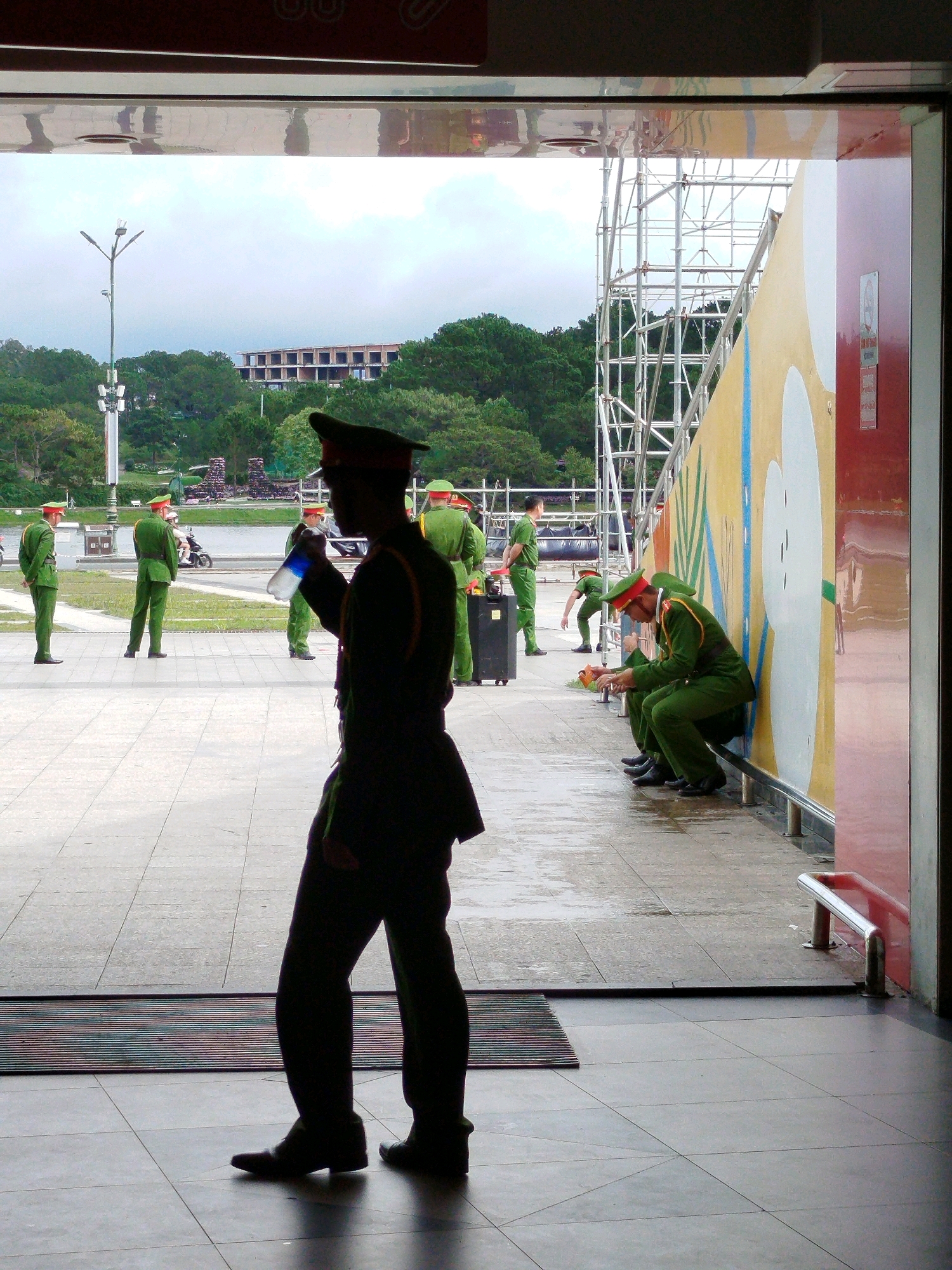 A group of men in green uniforms, likely police or security personnel, are visible in an outdoor covered area. In the foreground, a man stands in silhouette holding a water bottle. Further back, several other men in similar uniforms are either standing at attention, sitting against a wall, or interacting with a bag. The environment appears to be a tiled walkway with a red and white striped mat at the entrance. In the background, there is scaffolding, a brightly colored mural on a wall, trees, a building under construction, and a lamppost. The scene is likely in Da Lat, Vietnam, given the architectural style and vegetation.