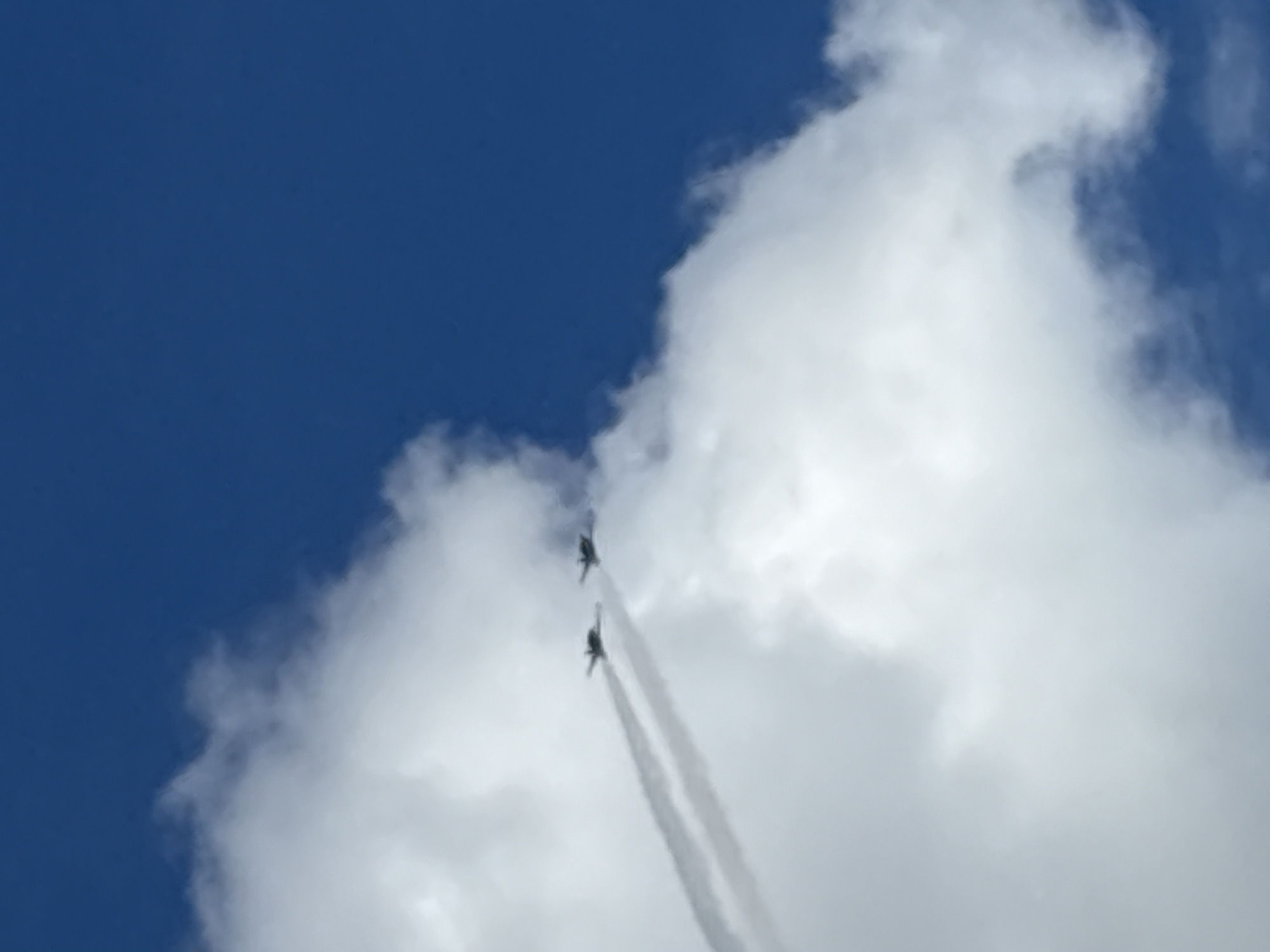 Two fighter jets fly through a bright blue sky, leaving trails of white smoke behind them. The jets are positioned at different altitudes and angles relative to each other, with the leading jet slightly higher and to the left of the trailing jet. They are flying over a dense layer of white clouds, which obscures the ground below. The image captures the dynamic movement of the aircraft against the backdrop of the sky and clouds. The location is identified as Odrava, Czechia.