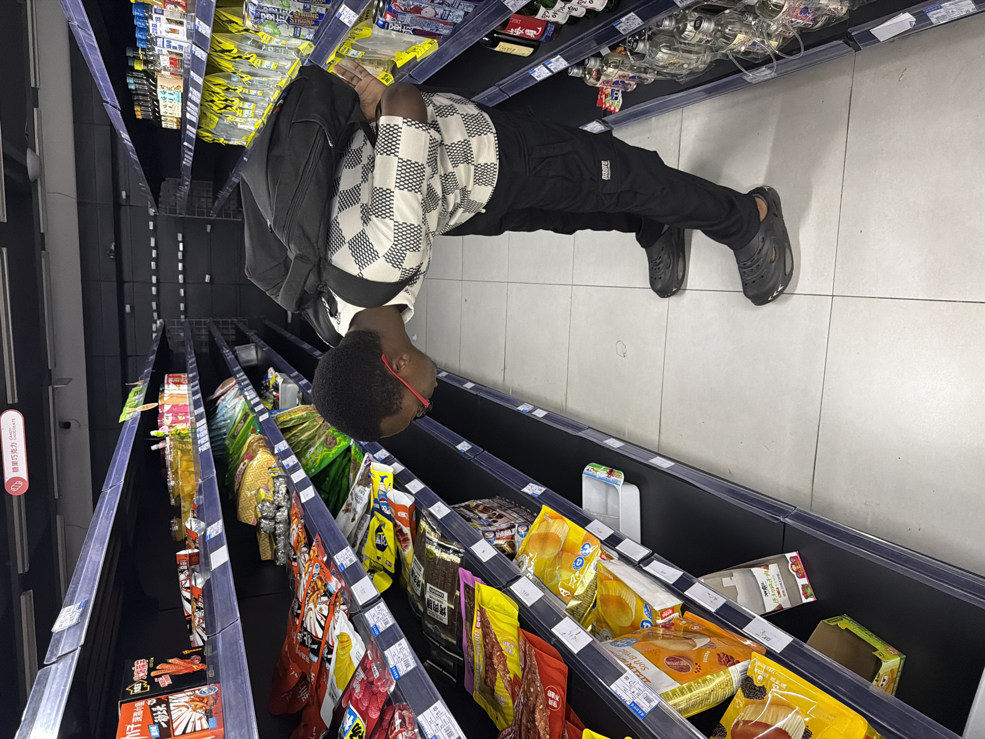 A young Black male is viewed from behind and at an angle, wearing a black backpack, a black and white checkered long-sleeved shirt, black cargo pants with a white logo on the left pocket, and black clogs. He is in a grocery store aisle, bending over to look at a shelf stocked with various packaged snacks. The shelves on the left are filled with brightly colored snack bags, while the shelves on the right contain bottles of alcoholic beverages. The flooring is tiled in a light beige color. The overall environment suggests a modern supermarket. The image location is the city of Jiangjin, China.