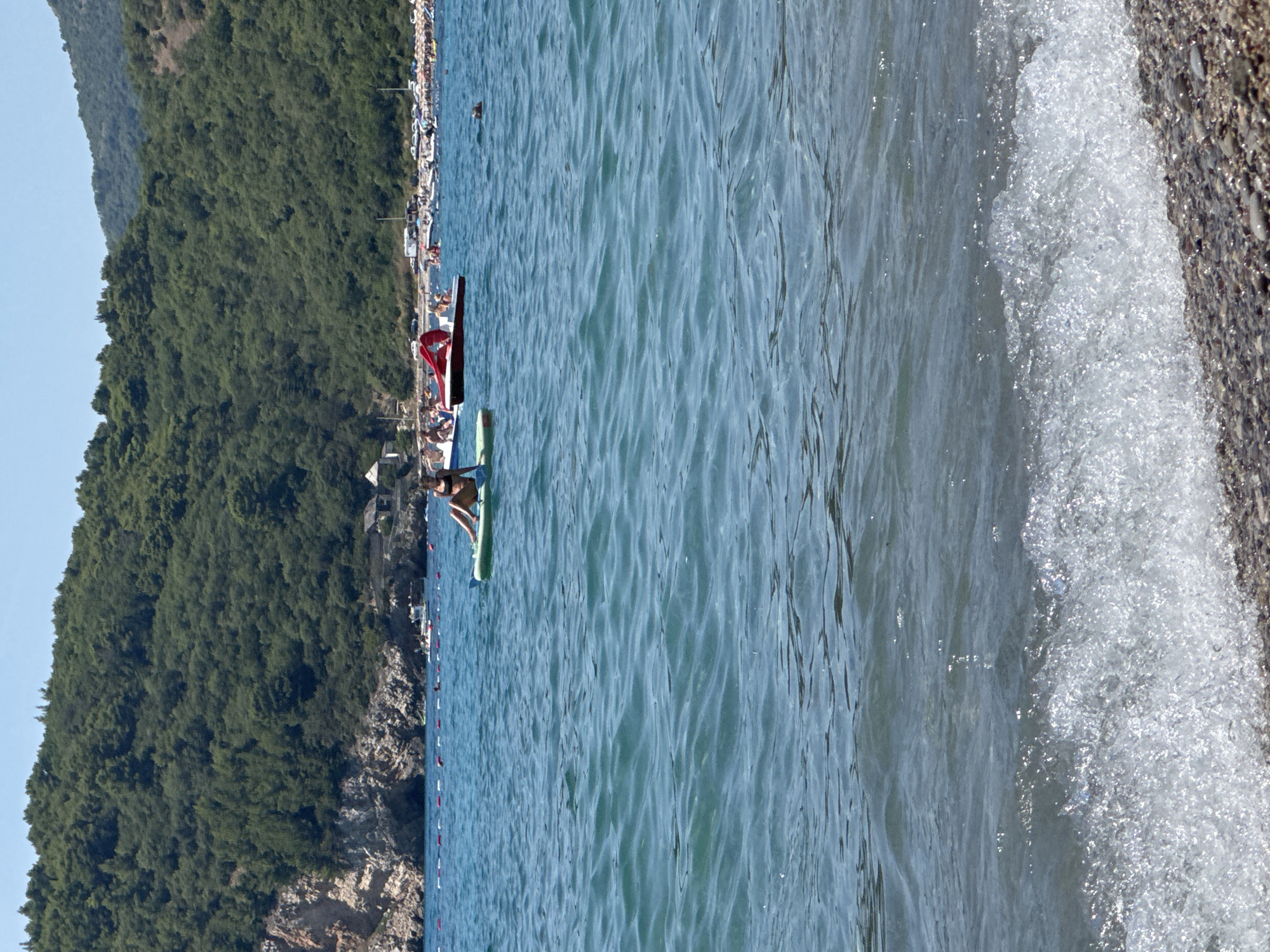 In the city of Đurovići, Montenegro, a bright, sunlit day showcases a serene coastal scene. The azure waters of the Adriatic Sea stretch towards a verdant, tree-covered mountainside that forms the backdrop. In the foreground, gentle waves lap against a pebble beach.
Further out in the water, a woman in a black bikini is seen relaxing on a green paddleboard. To her left, a red pedal boat with a canopy is occupied by several people. In the distance, other boats and numerous beachgoers dot the shoreline, suggesting a popular summer destination. The water is calm with slight ripples, and a string of buoys marks a boundary in the sea. The overall impression is one of a leisurely summer day at a picturesque European beach.