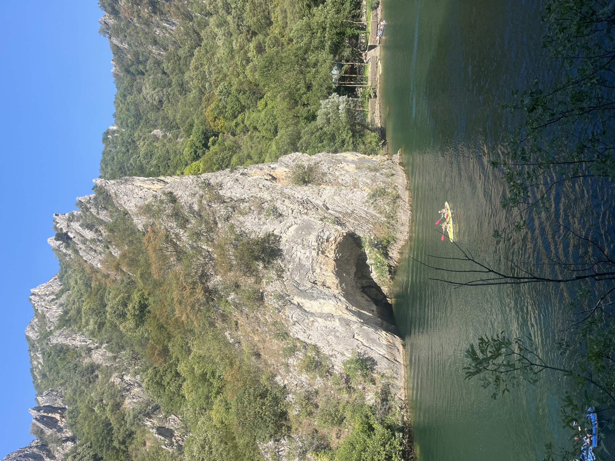 In a remote, mountainous region with steep, tree-covered slopes, a wide, deep green river flows. The rocky cliffs are weathered and appear ancient. A prominent cliff face juts out over the water, revealing a large cave opening. On the right bank, a small, rustic wooden structure sits nestled among trees, with a few people gathered on steps leading to the water. Several kayaks and inflatable boats are visible on the river. In the foreground, a bright yellow kayak with two people paddling glides across the water, while two blue inflatable boats are seen in the lower left corner of the frame. The sky is a clear, bright blue.