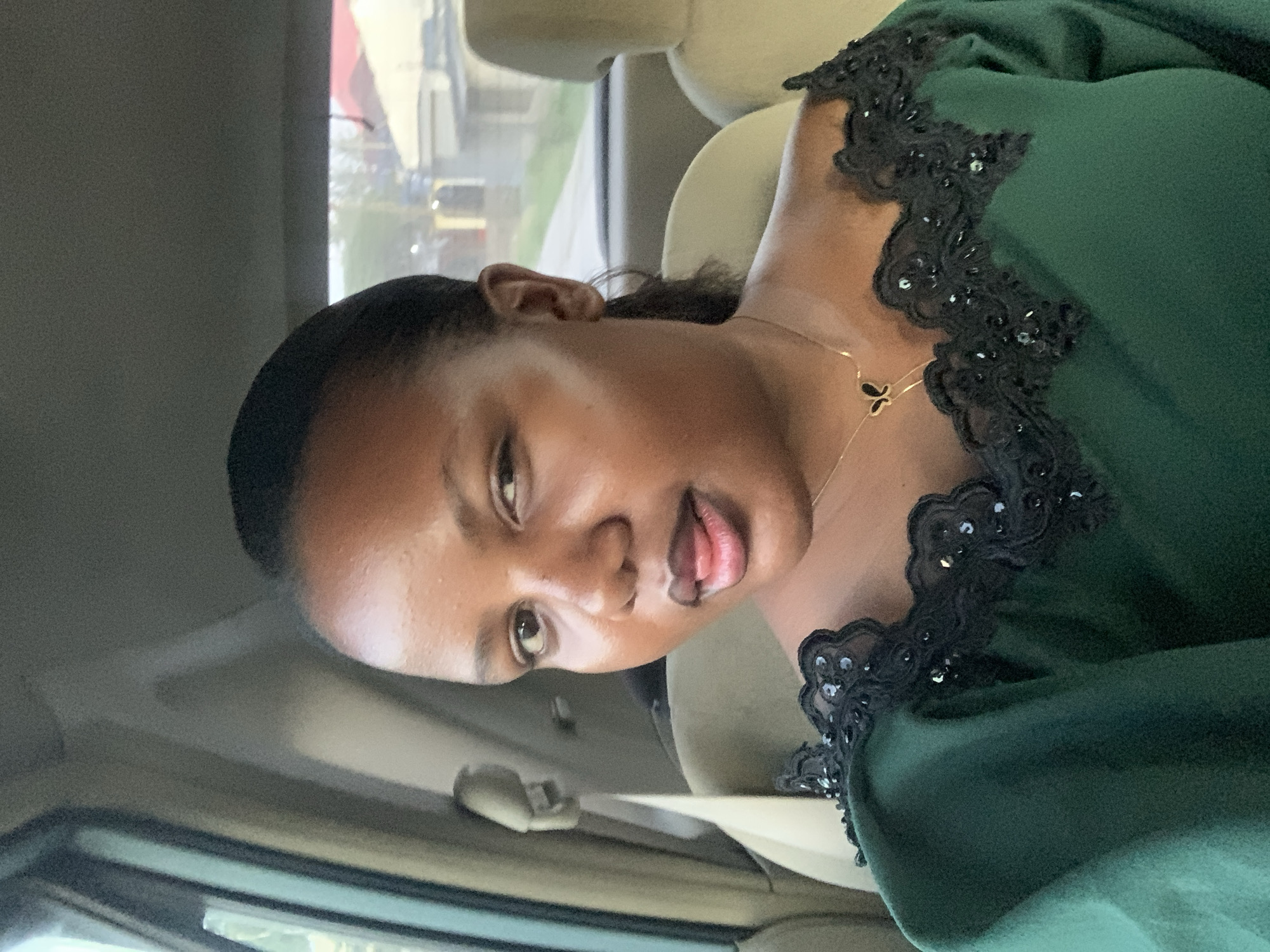 A medium shot of a dark-skinned woman in a green dress with black lace trim. She is wearing a gold necklace with a butterfly pendant. She is looking at the camera with her tongue slightly sticking out. The woman is inside of a car. The background shows an out-of-focus cityscape. The image was taken in Dar es-Salaam, Tanzania.