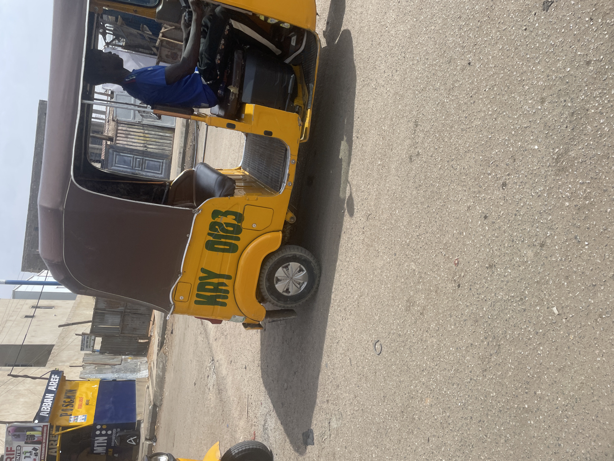 The image shows a yellow motorized tricycle, also known as a "keke", on a paved road in an urban environment. The keke has the text "KRY 0123" painted on its side. A person wearing a blue shirt is driving the keke. In the background, there are buildings and structures, including one with visible advertising signs for "ABBAN AREF", "POSEMIN", and "MTN". The location of the scene is in the city of Unknown, Unknown.