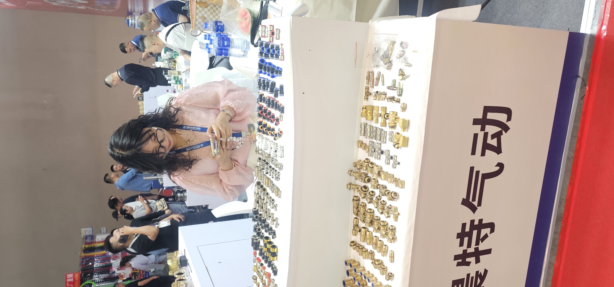The image shows a woman with long dark curly hair and glasses, wearing a pink blouse and a lanyard, standing at a table filled with various small metal connectors. She appears to be examining or assembling some of the connectors. Other individuals can be seen in the background, some looking at the display on the table. The table has a white backdrop with Chinese characters in dark print on the right side. There are bottles of water and other items on the table. The location appears to be an exhibition hall or trade show, potentially in Liangjiang, China.