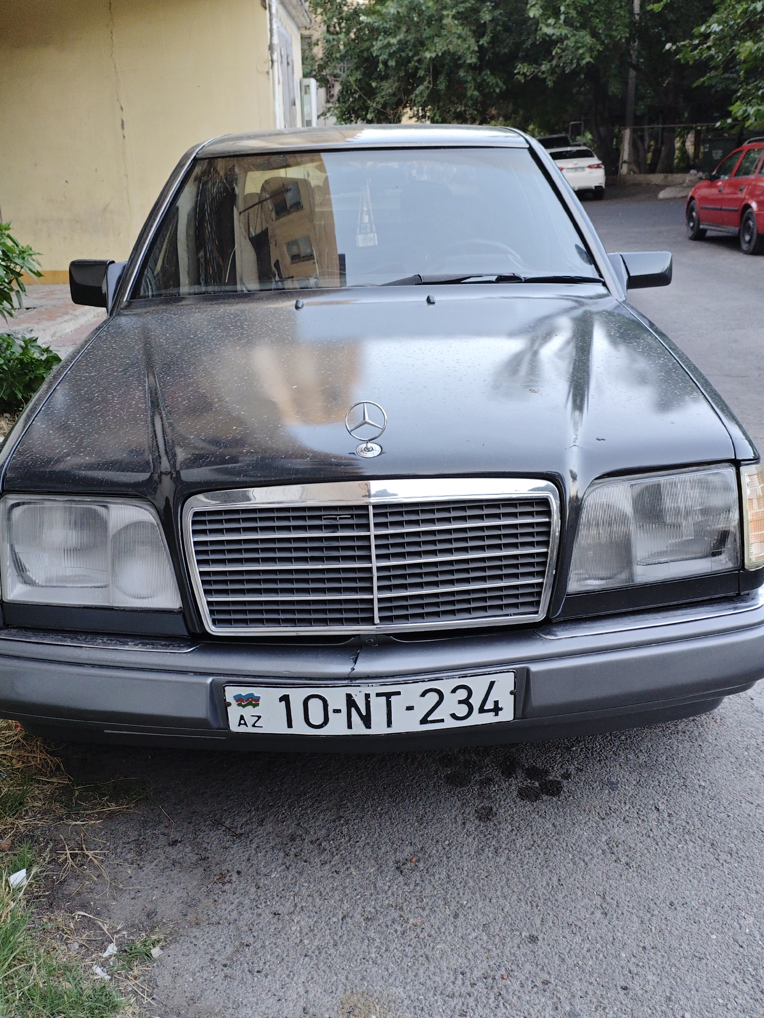 The media file displays a front view of a dark-colored Mercedes-Benz sedan parked on a street. The car has a prominent chrome grille with the Mercedes-Benz star logo at the top center of the hood. The headlights are clear, and the car has a license plate that reads "AZ 10-NT-234". The license plate also features the flag of Azerbaijan.
The car is parked on an asphalt road, with a sidewalk and some grass to the left. In the background, a light yellow building with windows is visible, and lush green trees line the street. Other vehicles are present in the distance, including a red car on the right and a white car behind the Mercedes.
The scene appears to be set during the daytime, with overcast or shaded lighting, suggesting it might be a cloudy day or the area is in shadow. There are no people visible in the frame. The overall impression is of a parked vehicle in an urban environment.