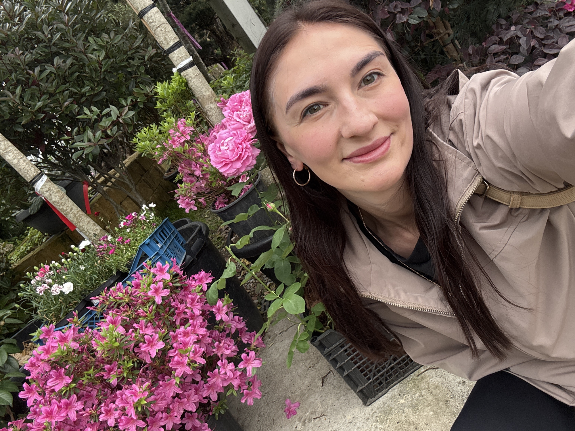 A woman stands in a garden setting, likely in the city of Sochi, Russia. The scene is filled with plants and flowers, notably pink azaleas and roses. The woman is fair-skinned with dark hair, wearing a light-colored jacket and gold hoop earrings. She is smiling and facing the camera. Behind her, there are various plants, trees, pots, and a blue crate. A portion of a white pole or tree trunk wrapped in black material is visible in the background. The ground appears to be concrete.