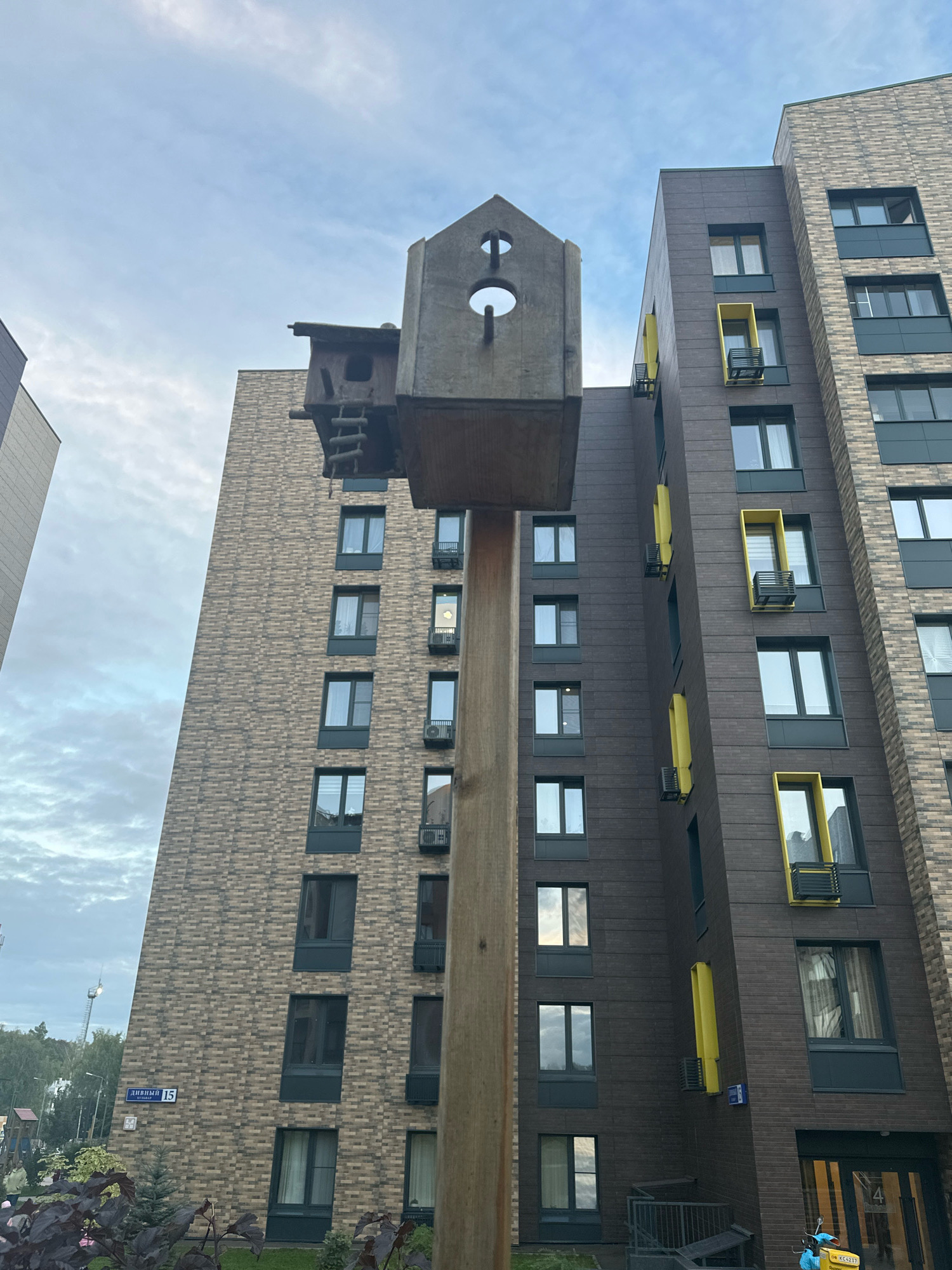 The image is an outdoor, low-angle shot of a modern apartment building with a unique birdhouse structure in the foreground. The apartment building on the right is dark gray with intermittent bright yellow window frames and balconies. The building on the left is covered in light brown brick. The birdhouse, made of wood and concrete, is mounted on a tall wooden post and has multiple levels and openings. The sky is overcast with some visible clouds. A street sign that reads "ДИВНЫЙ 15" (Divnyy 15) is visible on the brick building. In the lower left, some dark foliage is in focus. The overall scene appears to be in a residential area.