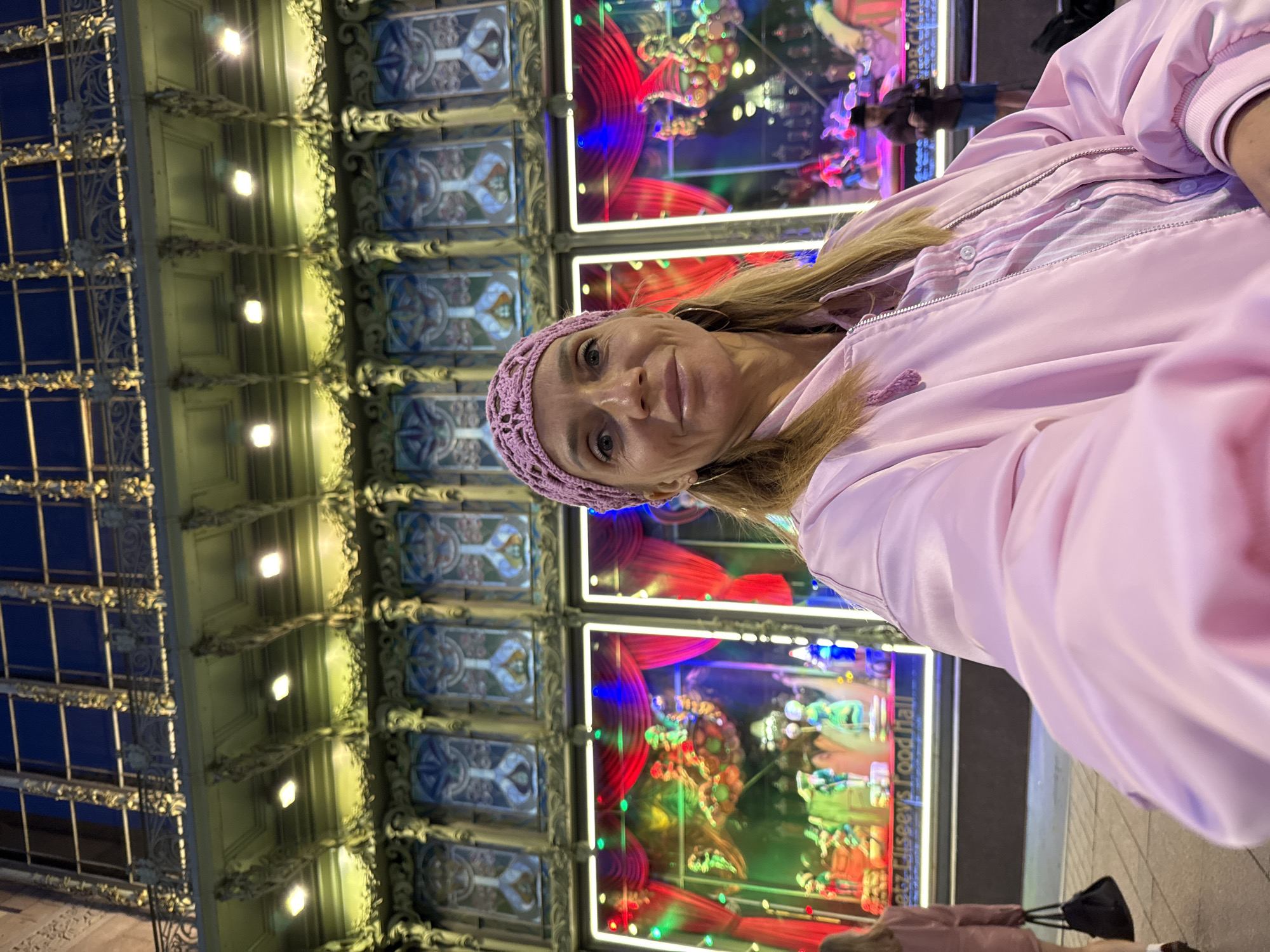 The image shows a woman in the foreground, taking a selfie outdoors at night in Saint Petersburg, Russia. She is wearing a pink knitted headscarf and a pink bomber jacket over a plaid shirt. Behind her, the facade of a building is visible, adorned with ornate architectural details and lit windows. The windows appear to be displaying elaborate dioramas or scenes, possibly shop displays or theatrical decorations, with red curtains and colorful lighting. In the background, other people are visible, as well as streetlights. The architecture suggests a historic or ornate part of the city. The text on one of the windows reads "atrz Eliseyys Food Hall".