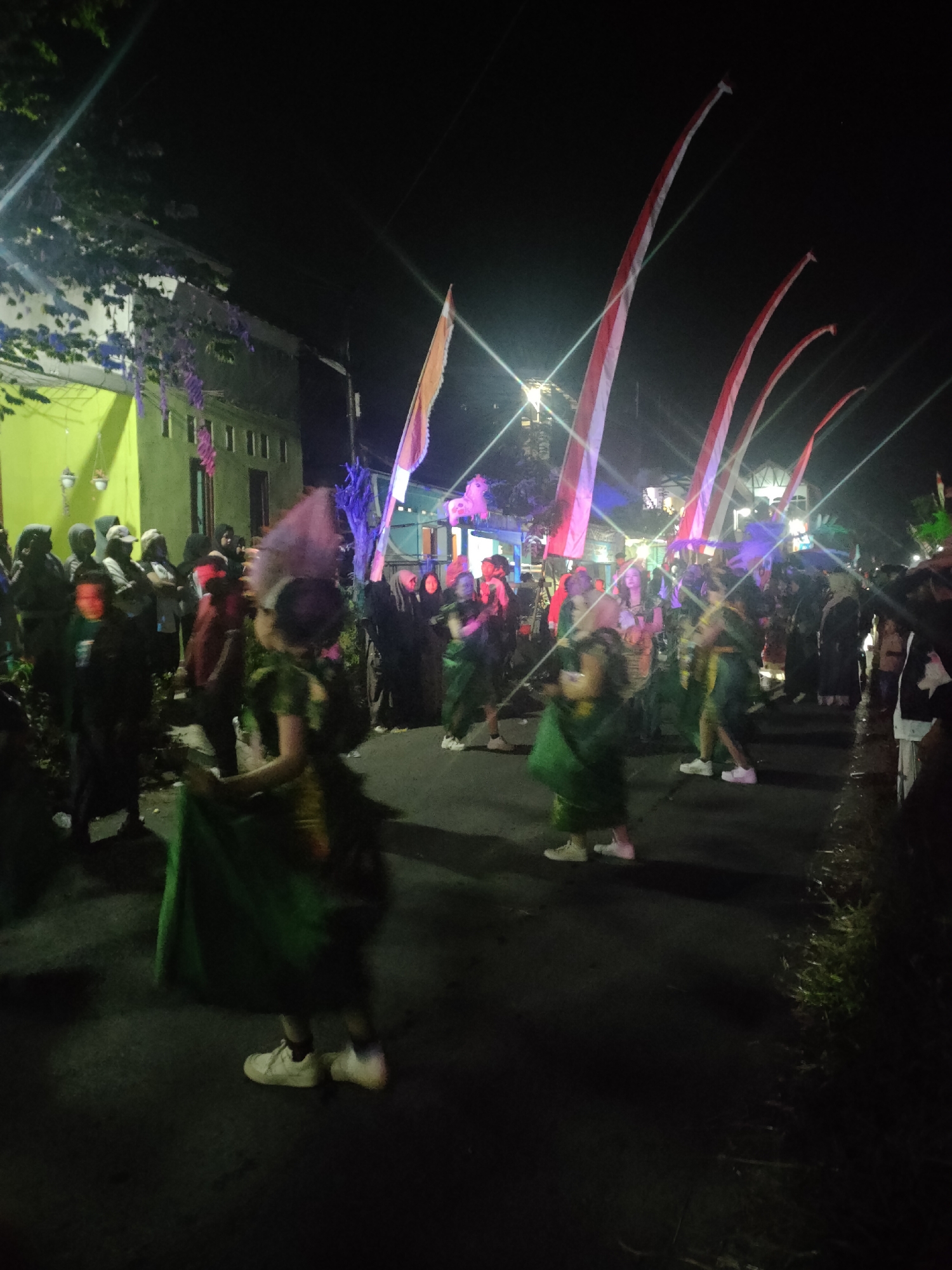 The image depicts a nighttime street scene in Tongaswetan, Indonesia, likely during a cultural event or festival. Several individuals, some dressed in green costumes with what appear to be ceremonial headdresses, are walking or dancing down a street. Tall, red and white striped flags are prominently displayed, likely Indonesian flags or decorative banners, extending into the dark sky. Buildings with yellow and light-colored facades are visible on the left side of the street, illuminated by artificial lighting. The ground appears to be asphalt, with grass or vegetation along the right side of the road. The overall atmosphere suggests a lively and festive gathering.