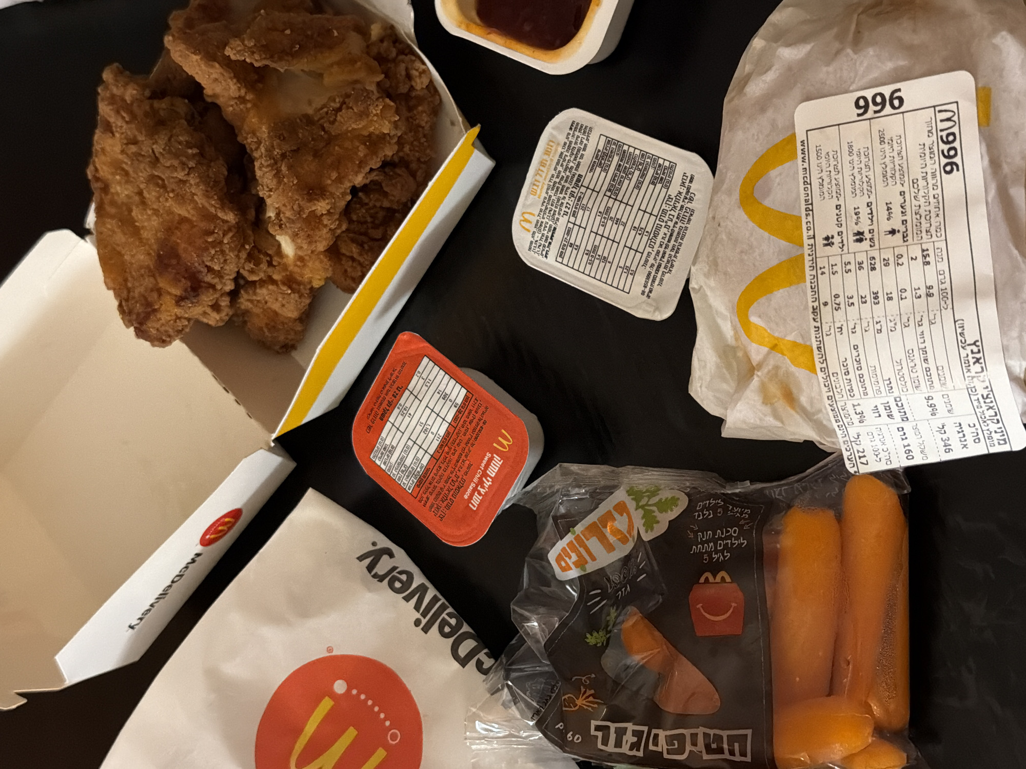 This media file captures a McDonald's takeout or delivery meal laid out on a dark surface, indicative of an indoor setting in Tel-Aviv, Israel.
The main subjects are various McDonald's food items and packaging. A white cardboard box on the left contains several pieces of golden-brown fried chicken or chicken nuggets. Next to it, a white McDonald's "McDelivery" bag is visible, partially obscured. A clear plastic bag of baby carrots, labeled "טיפי גזר" (Tip-carrot / Baby Carrot) in Hebrew, sits in the lower right, with text indicating it's "for children under 5".
Several sauce containers are present: one red container prominently labeled "Sweet Chili Sauce" in both English and Hebrew, and a white container with extensive nutritional information in Hebrew, likely a honey mustard or ranch sauce. A crumpled paper wrapper, bearing the McDonald's logo and the number "996", also displays nutritional facts and text in Hebrew, including "מיני קראנצ'י ראנץ" (Mini Crunchy Ranch) and a recommendation to "eat immediately."
Visible text includes the "McDelivery" logo, "www.mcdonalds.co.il", product codes like "996" and "M996W", and detailed Hebrew nutritional information and kashrut certification ("כשר פרווה בהשגחת וועד הכשרות פורטוגל" - Kosher Parve under the supervision of the Portugal Kashrut Committee). Importer information for "Aloniel Ltd., Beer Tuvia" and manufacturer "Mendes Gonsalves, Portugal" are also discernible on sauce containers. The scene suggests a recently arrived or unpacked meal.