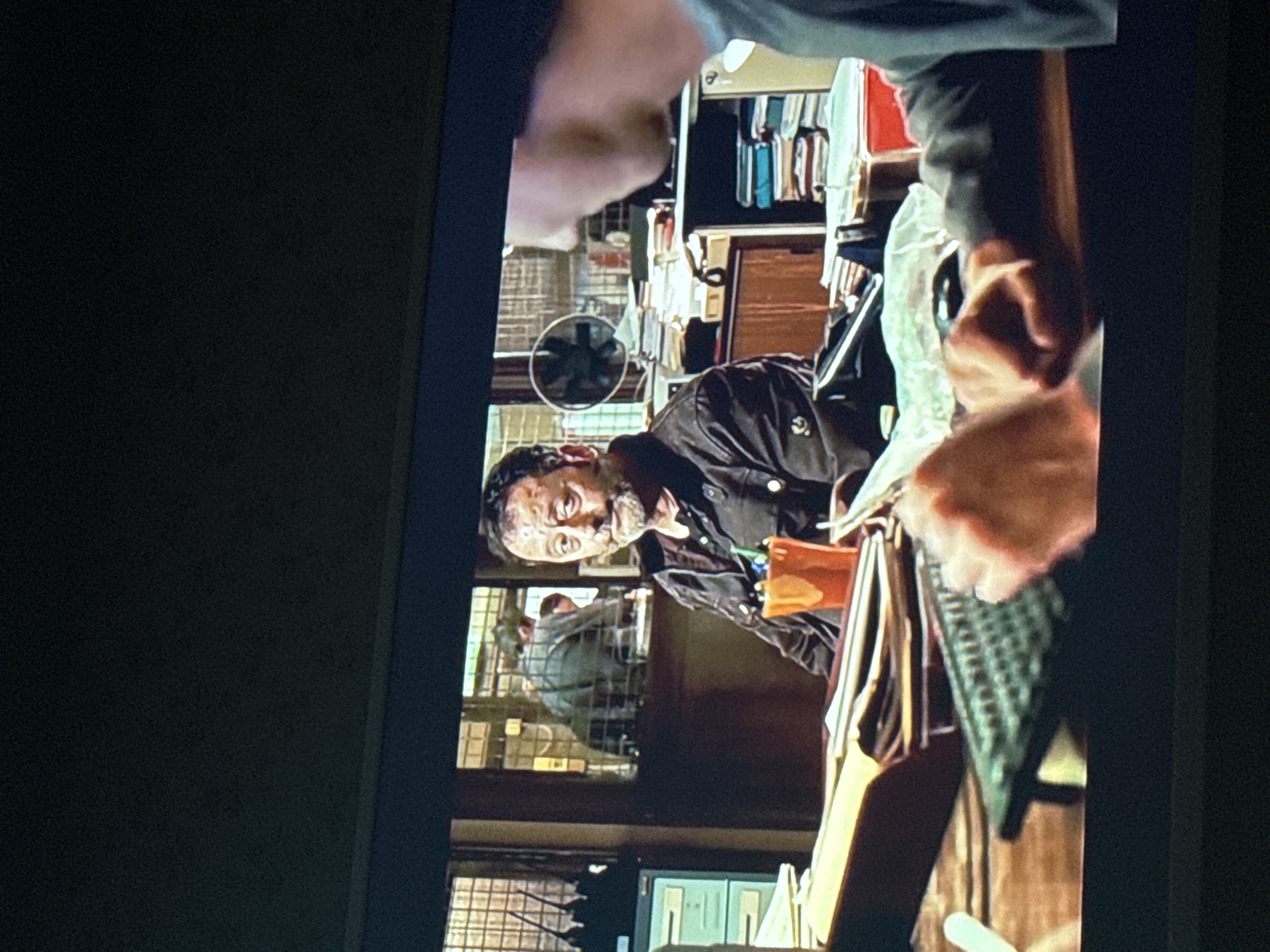 The image shows a man with a beard, wearing a dark button-down shirt, looking at the viewer. He is inside a cluttered office. Behind him is another person in a grey hoodie behind a metal grate. A small fan sits on a shelf with various objects and books. In the foreground, a hand holds a grey metal grate. It's impossible to pinpoint the exact location in Mytishchi, Russia, without further information.