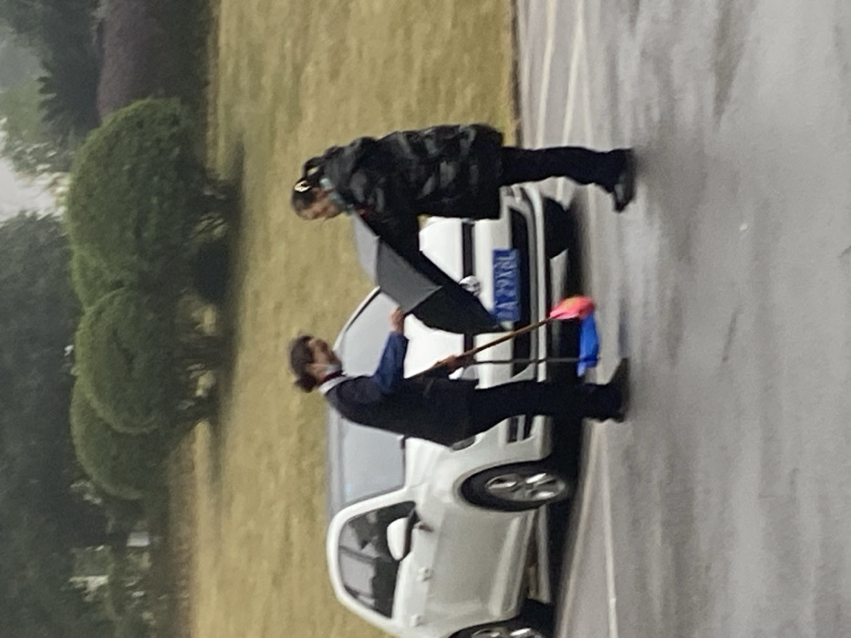 Two individuals are standing next to a white car, parked on a paved surface near a grassy area with shrubs. One person holds an umbrella over the car. Another individual stands near the front of the car, holding a stick with a colorful object attached to the end. The car has a license plate visible. The setting appears to be outdoors on a cloudy day.