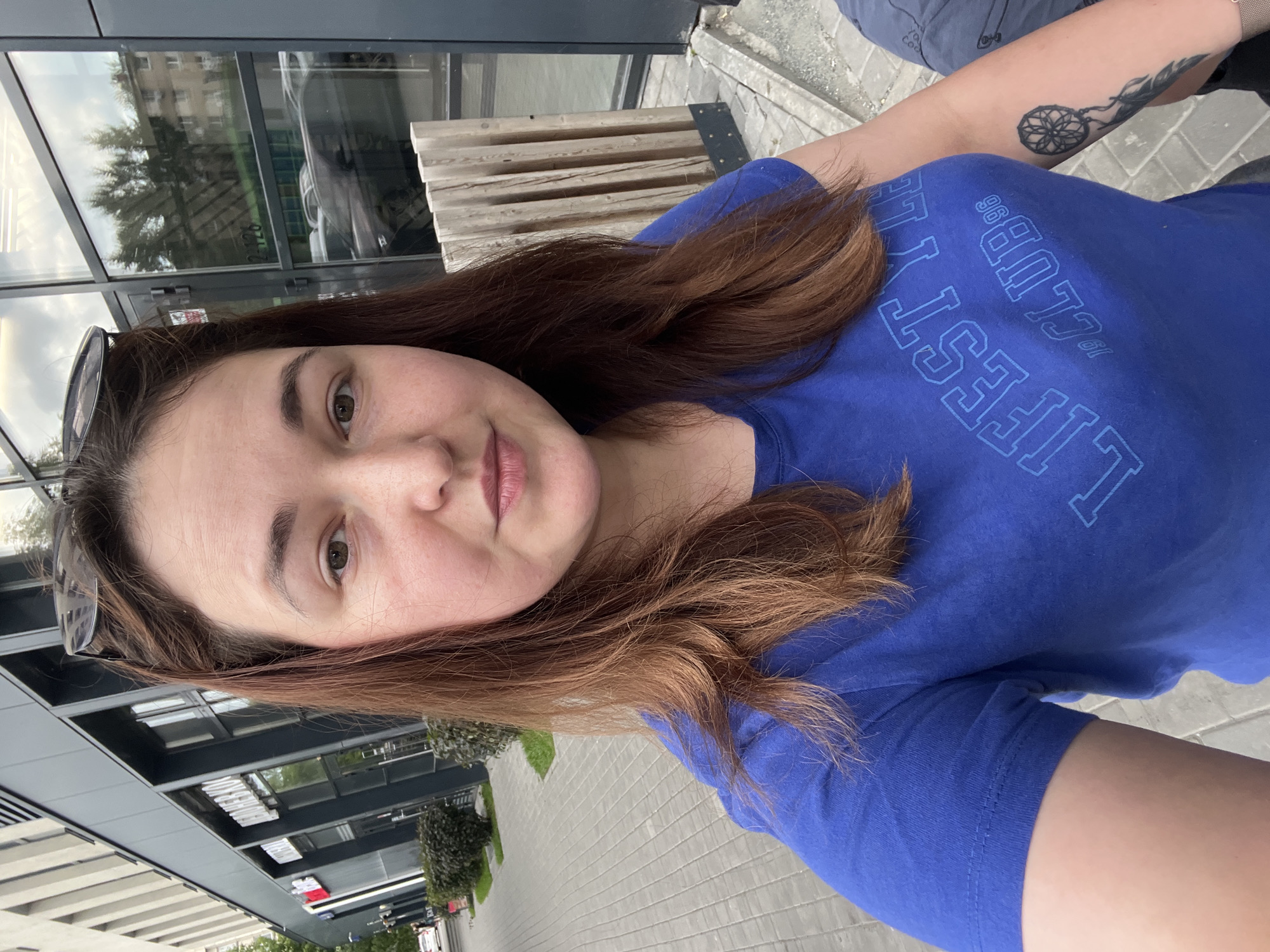 A woman is taking a selfie outdoors. She has long, brown hair and is wearing a blue t-shirt with blue text that reads "LIFESET CLUB" and "98". She is also wearing sunglasses pushed up on her head. In the background, there is a modern building with large windows, some trees, and a paved walkway. A wooden bench is visible to the right of the woman. The scene appears to be during the daytime, with overcast or cloudy weather given the muted lighting and reflections in the windows. The woman's expression is neutral. The location is identified as Chelyabinsk, Russia.