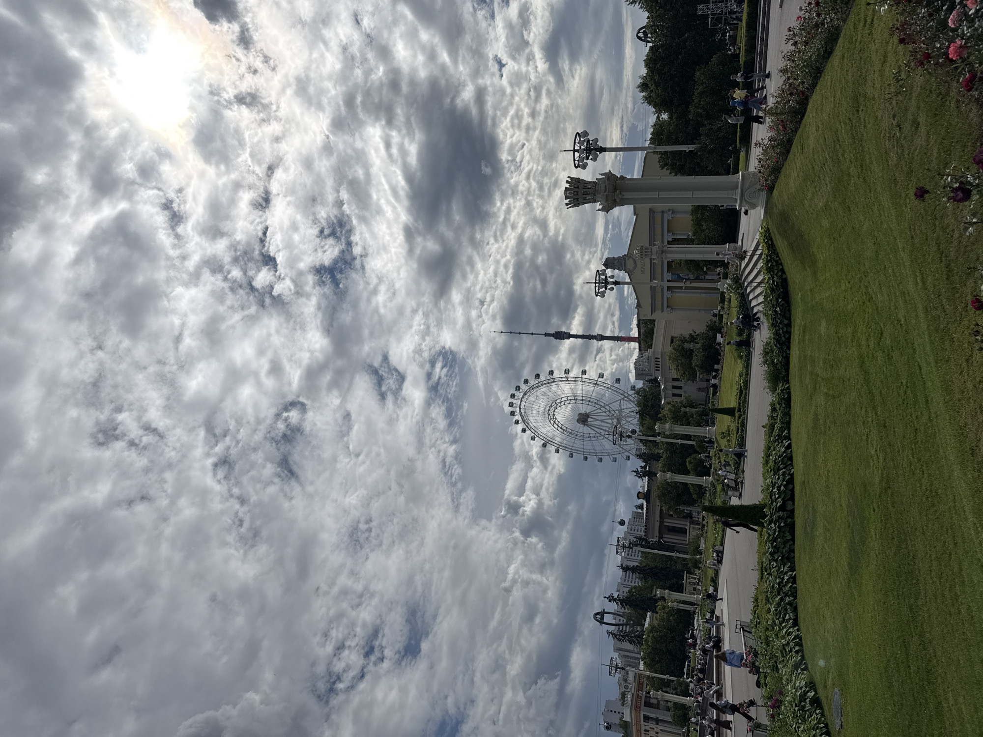 This is a wide-angle, slightly elevated shot of a park in Moscow, Russia, likely VDNKh (Exhibition of Achievements of National Economy), given the presence of the iconic Ferris wheel and a tall tower in the background. The sky is dramatic with a mix of dark and light clouds, with the sun breaking through on the right side.
In the mid-ground, a large, white Ferris wheel is prominently featured to the left of center. To its right, a tall, slender radio tower is visible. In front of the tower and Ferris wheel are several classical-style buildings with white columns and decorative elements. There are also several ornate lampposts.
The foreground is dominated by a lush green lawn with manicured flower beds along its edges. People are scattered throughout the park, walking, sitting on benches, and strolling. Some individuals are walking towards the buildings in the mid-ground, while others are visible in the distance. The overall atmosphere suggests a sunny, somewhat cloudy day in an urban park setting.