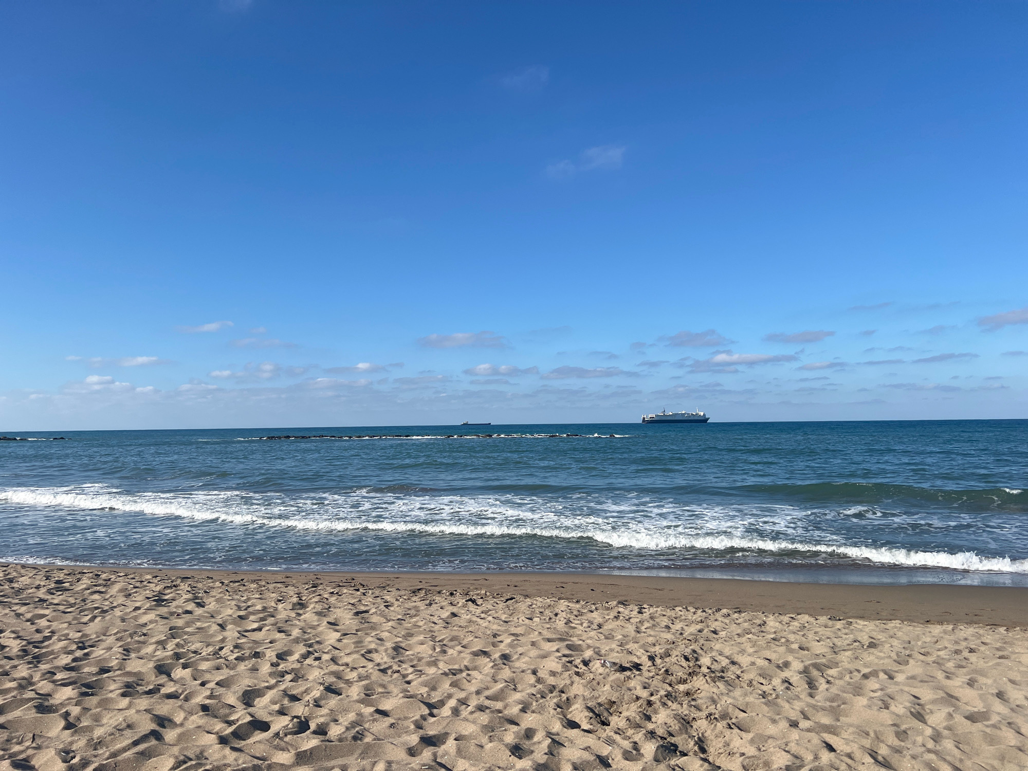 The image depicts a serene beach scene in Karasu, Turkey, under a clear blue sky with scattered white clouds. The foreground is dominated by textured sand with visible footprints. A gentle wave is breaking on the shore, creating a white foamy line. Beyond the shoreline, the sea extends to the horizon, with two cargo ships visible in the distance. One ship is closer to the left side of the frame, appearing smaller, while a larger, white cargo ship is positioned on the right side, closer to the horizon. The water is a deep blue, and the horizon line is sharp. The overall impression is one of tranquility and open space.