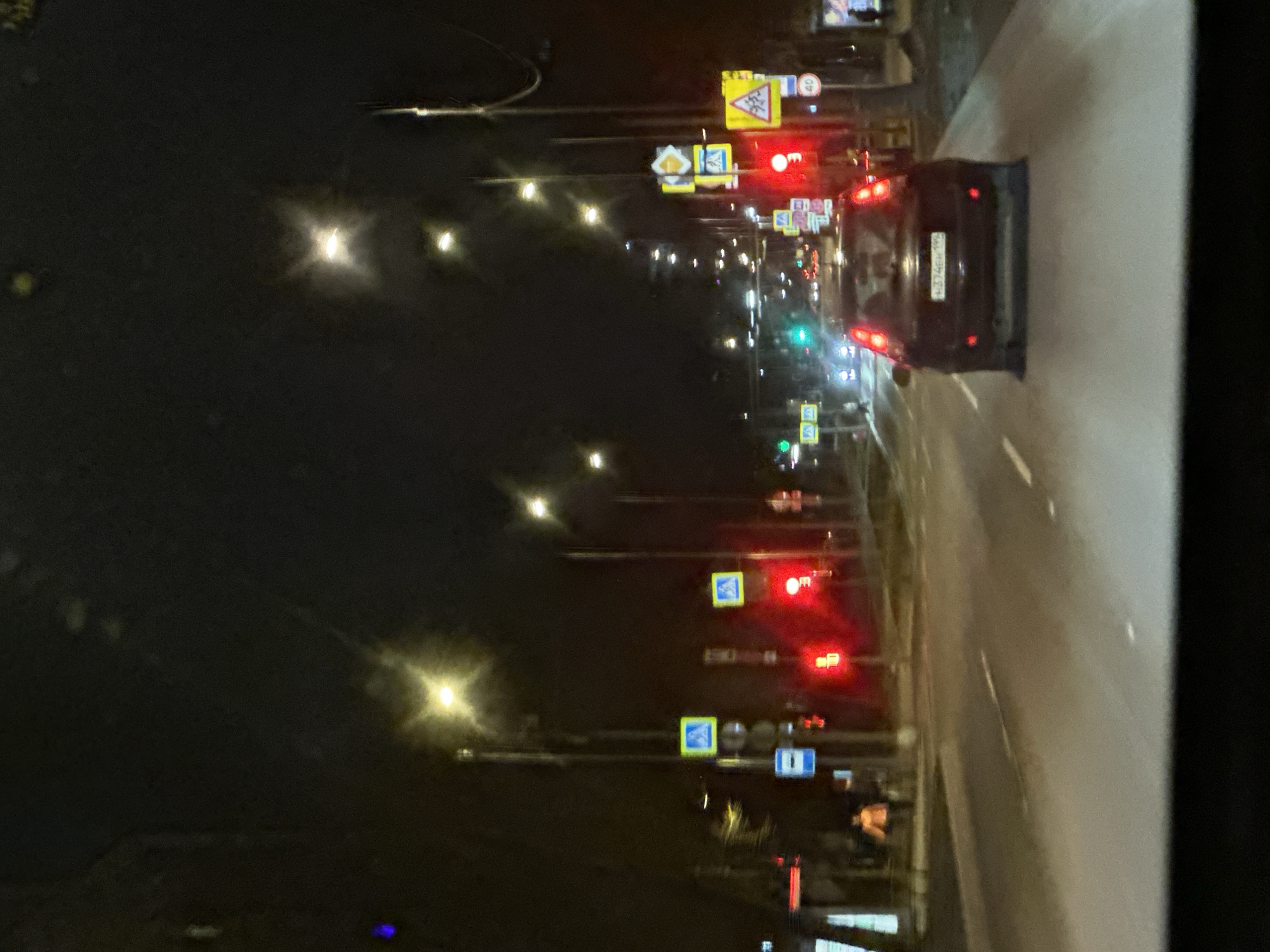 The image depicts a nighttime street scene in Moscow, Russia. A car with license plate number H674EH 197 is visible in the lane of traffic, stopped at a red light. Several traffic signals and street signs line the road, indicating intersections and pedestrian crossings. The overall environment is urban, illuminated by streetlights and vehicle headlights.