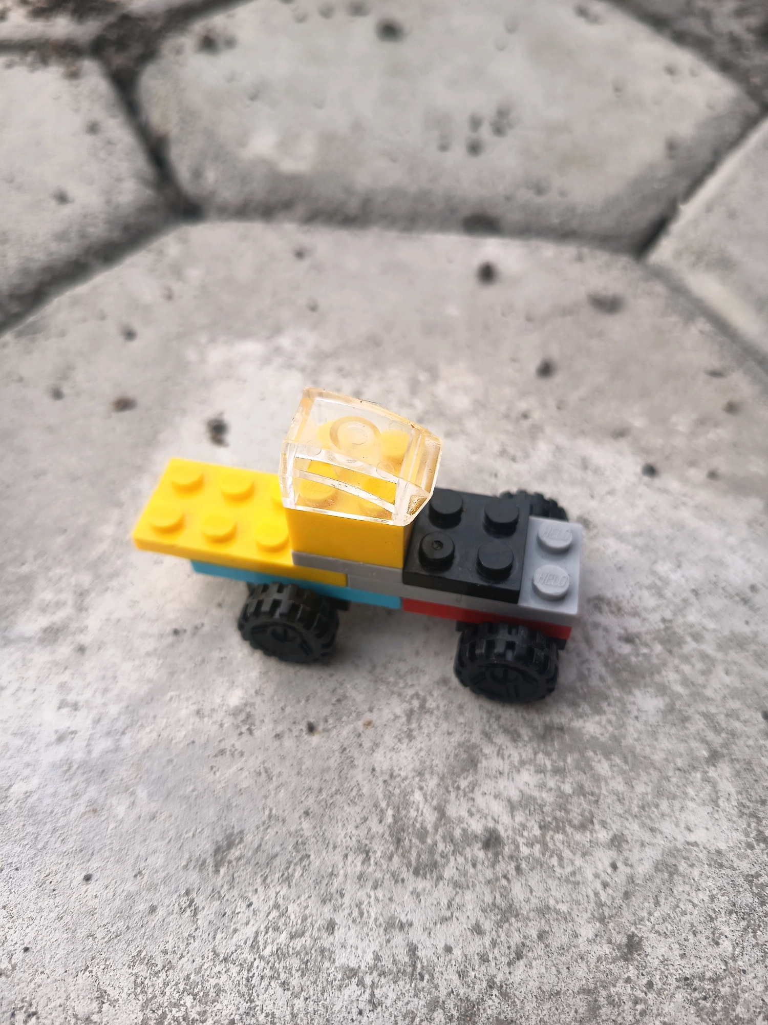A close-up, elevated shot shows a small, multi-colored toy truck made of Lego bricks resting on a textured, light gray concrete surface. The truck features a yellow base, a blue underside, black wheels, a red brick detail, and a transparent yellow Lego piece forming the cab. The concrete surface is composed of large, irregular hexagonal or pentagonal pavers, with visible dark lines between them. The lighting suggests an outdoor setting with soft shadows. The scene is devoid of people and activity, presenting a still-life composition of the toy. The location is specified as Sidorejo, Indonesia.