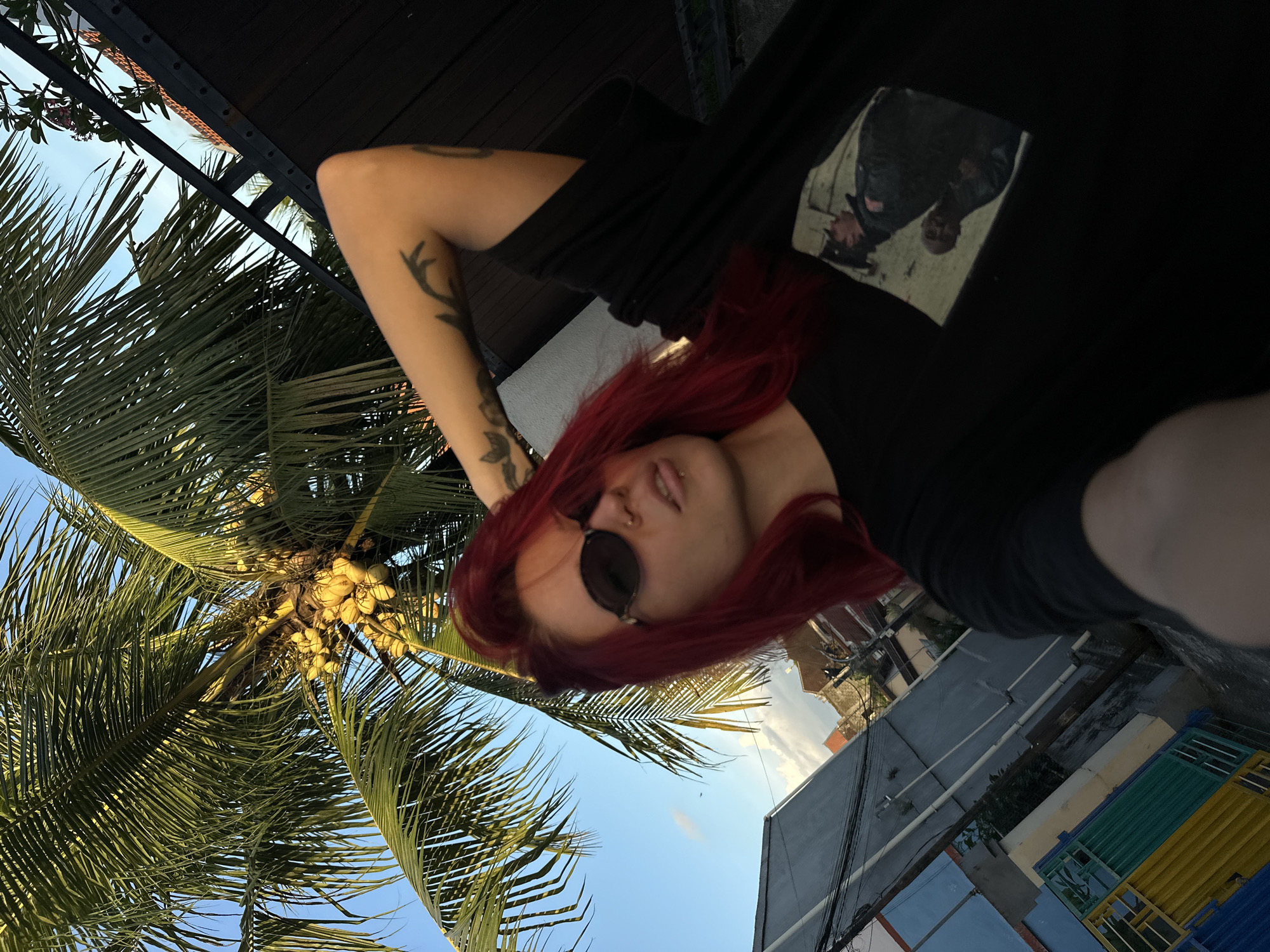 A low-angle shot of a person with red hair, wearing sunglasses and a black t-shirt with a print on it, with their arm raised and displaying tattoos. The person is positioned with a palm tree with coconuts on the left and buildings in the background. The location is likely North Kuta, Indonesia, based on the vegetation and building style.