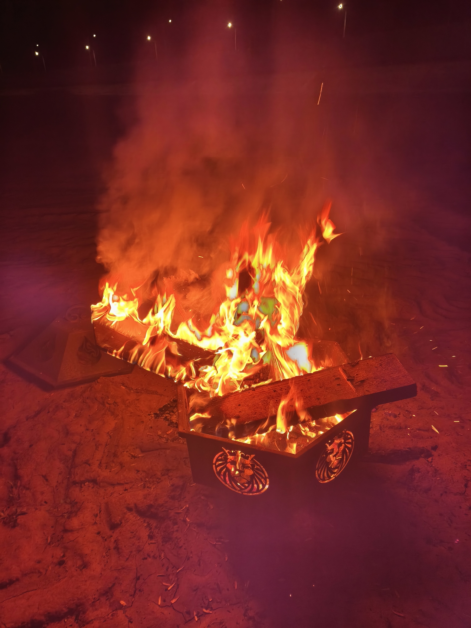 A rectangular metal fire pit, decorated with lion head cutouts, is filled with burning wood and flames. Smoke rises above the flames. The fire pit sits on a dirt or sandy surface. In the background, several distant lights are visible, likely streetlights. The location is Сокуры, Russia.