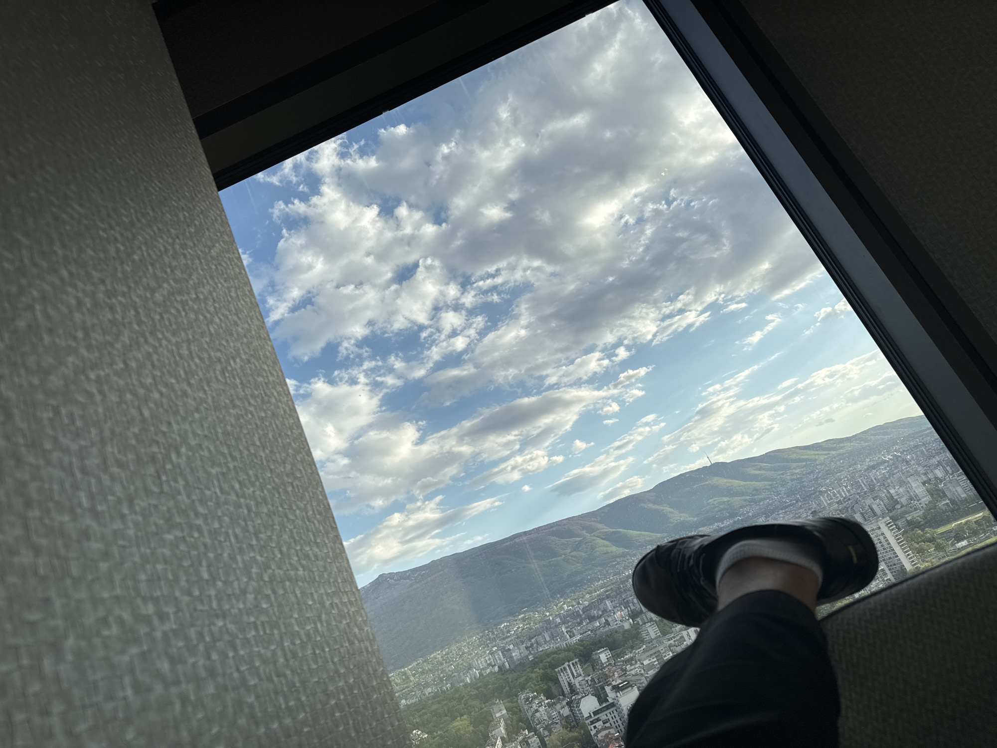 An indoor scene is captured from a high vantage point, likely from within a building looking out of a window. The camera's perspective is pointed towards the lower right corner of the sky, which is predominantly filled with blue interspersed with scattered white clouds.
The view through the window reveals a cityscape at the foot of a large, verdant mountain, which is clearly visible in the distance. Based on the visual evidence, the cityscape is identified as Sofia, Bulgaria.
Foreground elements include the textured surface of a wall or partition on the left side of the frame, and a partial view of a person's leg and foot resting on a surface near the window, suggesting a seated position. The person is wearing black pants and a black shoe with a white sock visible. The shoe is positioned in a way that suggests it is resting casually.
The lighting in the scene is bright, casting shadows on the wall and adding depth to the cloud formations. The overall mood is relaxed, with a clear emphasis on the contrast between the natural landscape and urban environment visible through the window.