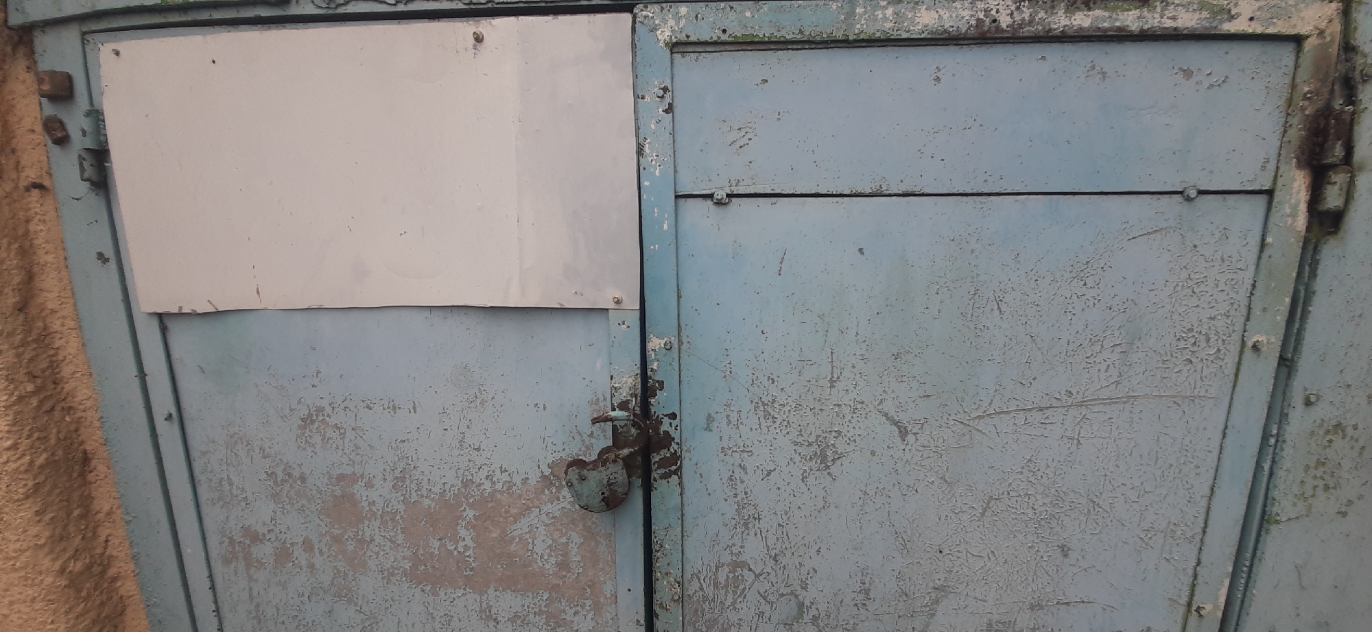 The image shows a close-up view of a weathered, light blue metal double door, likely part of a garage or shed. The surface of the doors is heavily chipped and peeling, revealing layers of rust and an underlying gray material. The left door has a rectangular light gray panel attached to its upper half, secured by small nails or screws. A rusty, heart-shaped padlock with a shackle is attached to the right door, near the vertical seam between the two doors.
The door is framed by a textured, fuzzy, light brown material on the far left, possibly a blanket or a rough fabric surface. The overall impression is one of age and disuse, with the peeling paint and rusted padlock suggesting exposure to the elements. There are no people visible in the image, nor any clear indication of the time of day or weather. The provided location context of Yessentukskaya, Russia, suggests an outdoor setting in a possibly rural or industrial area. No visible text is present in the image.