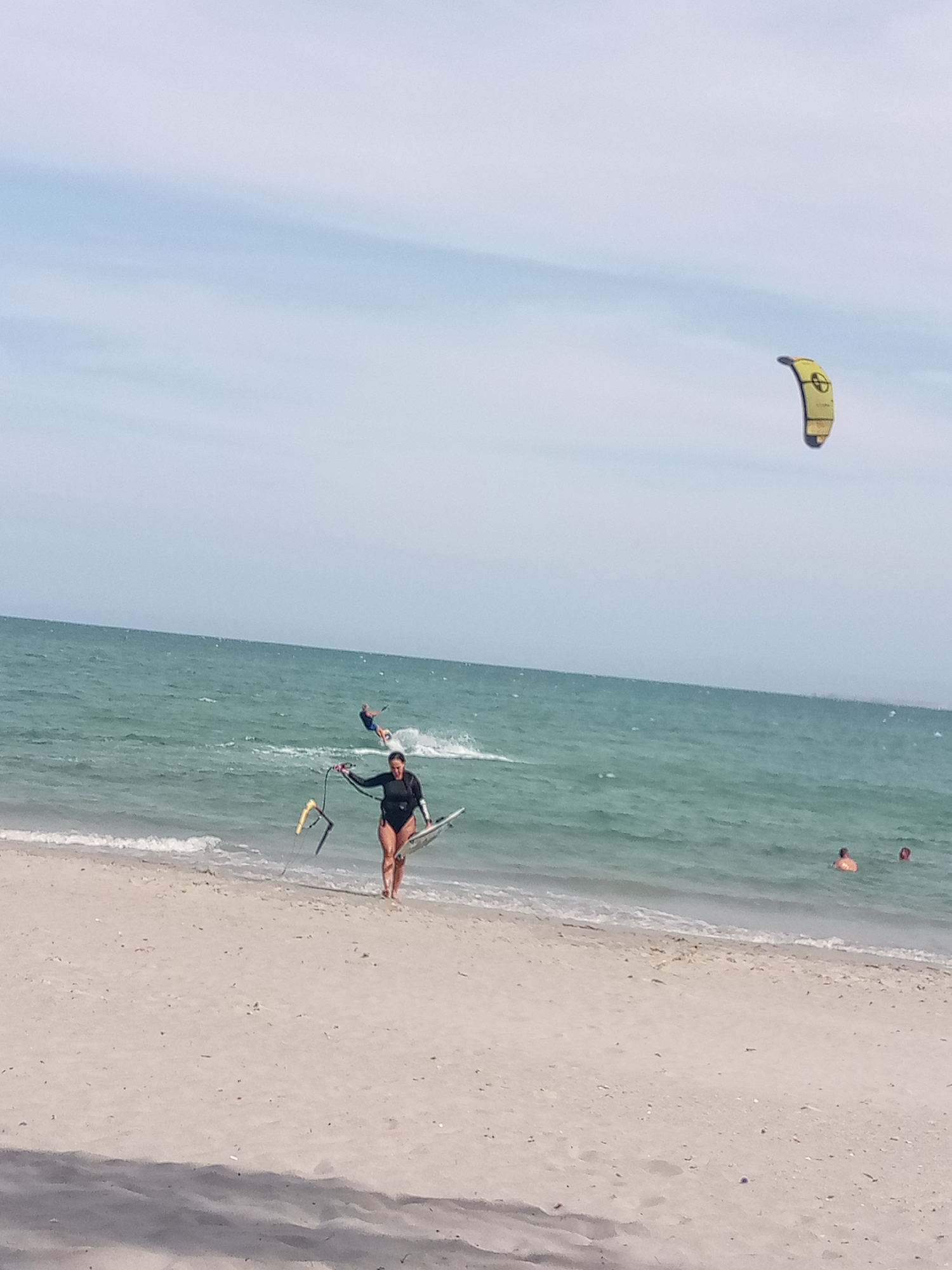 A woman in a black wetsuit stands on a sandy beach, holding a kiteboard and kite control bar, near the water's edge. In the water behind her, a person is kite surfing and two people are swimming. A yellow kite flies high in the sky. The location is El Yaque, Venezuela.