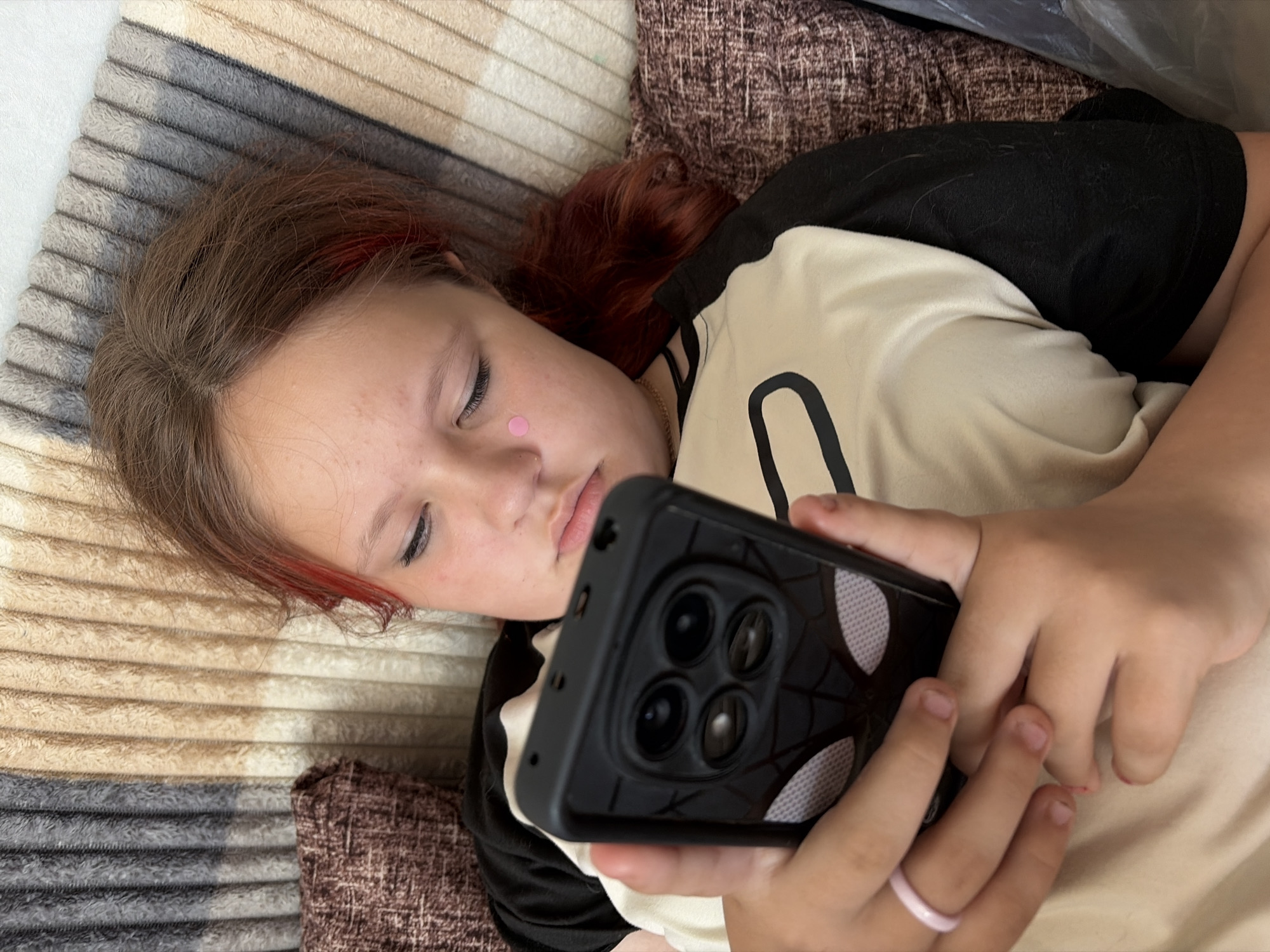A young person, presumably a child, is lying on a striped beige, brown and gray soft surface with a brown pillow in the upper right corner, possibly a sofa or bed. The subject has fair skin, light brown hair with reddish highlights, and is wearing a two-toned black and beige shirt. A small pink round object is adhered to their face, under the right eye. The subject is holding a black smartphone with a distinctive camera module and spider-web-like design on the back in their hands. The subject is looking at the smartphone, potentially engaging with it. A pink ring is visible on the small finger of the hand holding the smartphone. The location is likely indoors. There is no information to confirm that the location is in Chaikovsky, Russia.