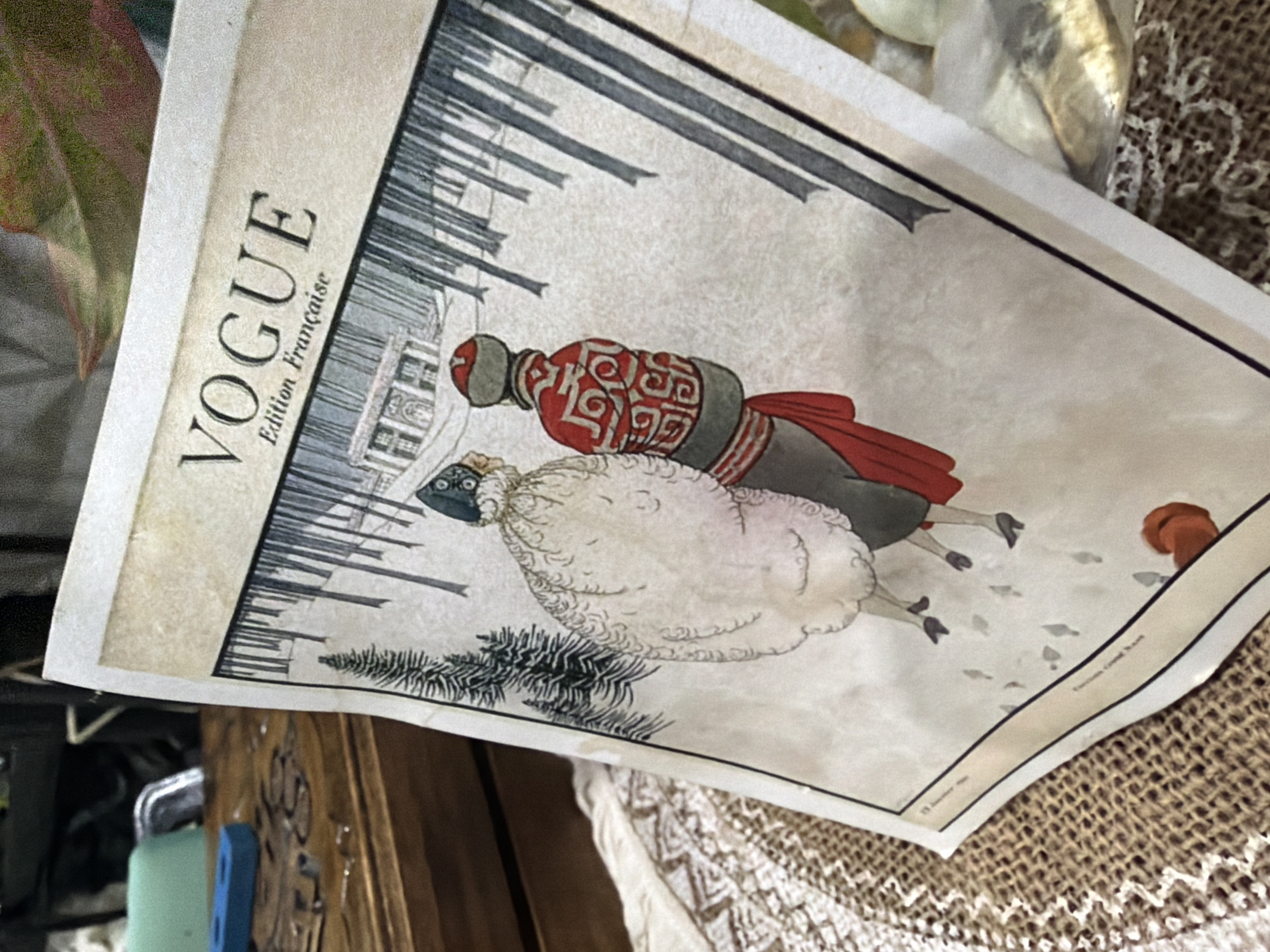 The image shows a close-up of a reproduction of a vintage Vogue magazine cover. The cover depicts a winter scene with two figures in period clothing walking through a snowy forest. The figures are viewed from behind, with one wearing a voluminous white fur coat and a dark hat, and the other in a red coat with intricate patterns and a fur-lined hat. In the background, a stately building can be seen amidst bare trees. The magazine is lying on a textured surface, possibly burlap, with a decorative lace cloth beneath it. To the left of the magazine, a wooden surface with carved details is visible, along with some blue and silver objects. The lighting suggests an indoor setting. The image does not depict any elements related to Baan Mae Nam, Thailand.