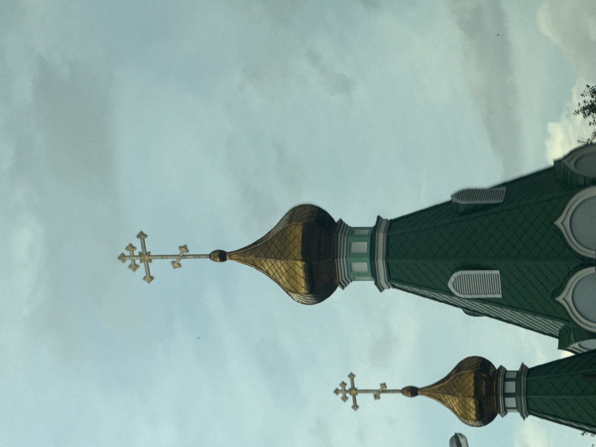 The image shows two onion domes of a church against a cloudy sky. Both domes are topped with golden crosses, with one cross being more ornate than the other. The larger dome, on the right, is part of a steeply pitched green roof, while the smaller dome, on the left, is also part of a green structure. The domes are golden and bulbous, characteristic of Russian Orthodox architecture. The sky is overcast with soft, diffused light. There are no people visible in the image. This scene is likely from Yekaterinburg, Russia, given the architectural style.