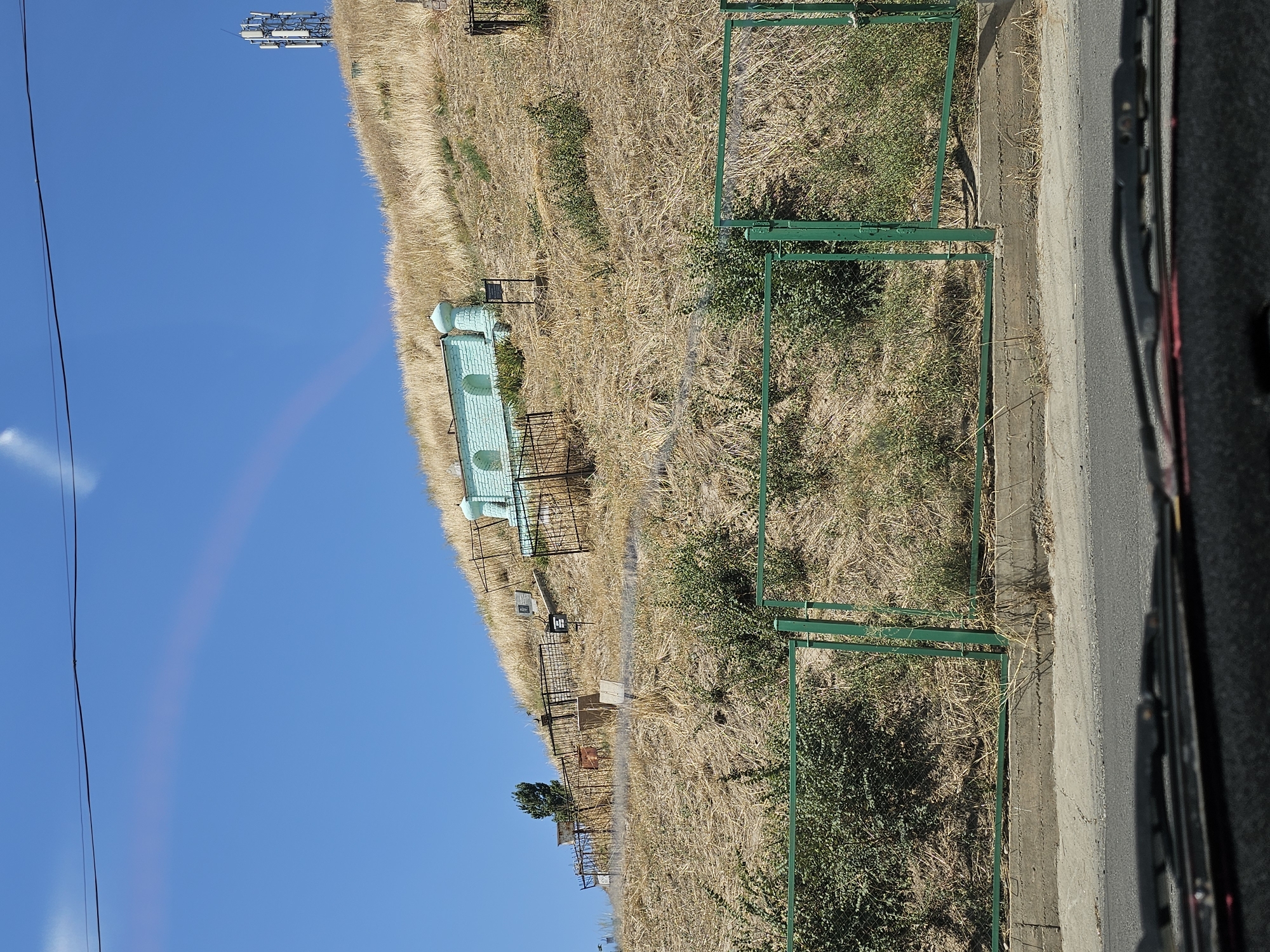 The image shows a view from a car looking up a dry, grassy hillside in the city of Özgön, Kyrgyzstan. At the top of the hill, there is a small, light blue structure with arched openings, possibly a mausoleum or shrine, surrounded by a fence. Scattered on the hillside are several gravestones and more fencing, indicating a cemetery. A modern cell tower is visible on the far right of the hilltop, and a faint rainbow is visible in the clear blue sky. A green metal fence runs across the middle of the image, separating the road and sidewalk in the foreground from the hillside above. The vegetation on the hillside is sparse and dry, typical of a warm, arid climate. The overall scene suggests a historical and religious site in a rural or semi-rural area.