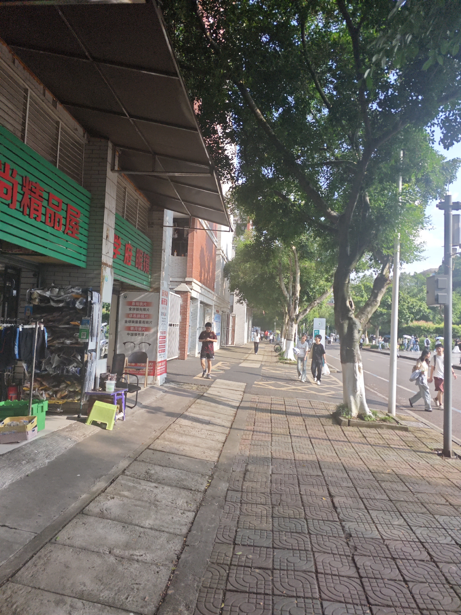 The image captures a street scene with a pedestrian sidewalk on the left and a road on the right. A building with a green awning and Chinese characters is visible on the left, with clothing racks and merchandise displayed outside. Further down the sidewalk, people are walking and browsing. Trees line the sidewalk and road, providing shade. The road has a traffic light pole and traffic. The overall setting suggests a commercial area within a city, consistent with the Chongqing High-tech Zone in China.