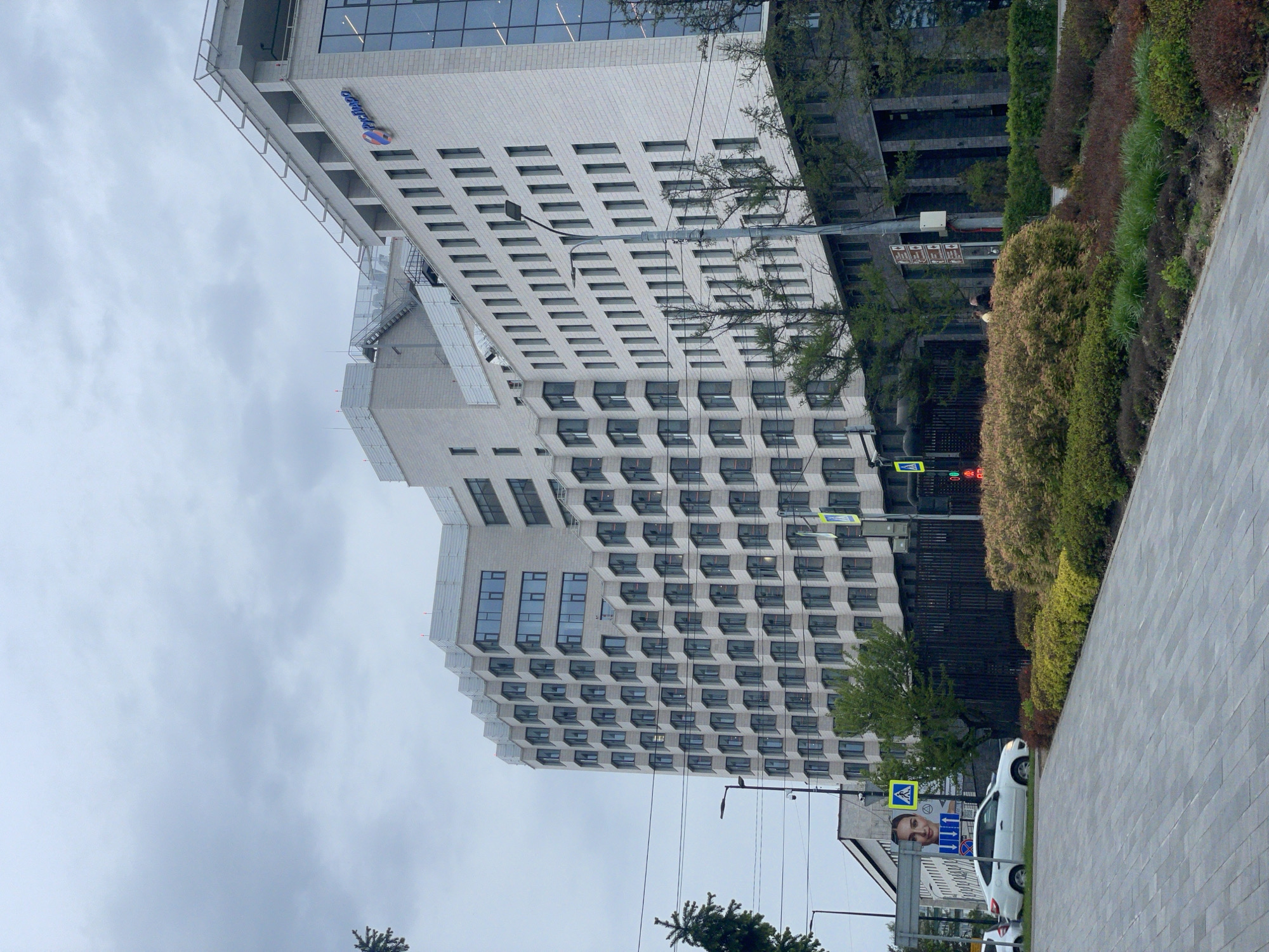 The image shows a high-rise building in Krasnoyarsk, Russia. The building has a white exterior with numerous windows. The Radisson Hotel logo is visible on the upper portion of the building. A pedestrian crosswalk sign is present, and a traffic light is also seen. A street with cars parked along it runs parallel to the building. Bushes and plants are present in the foreground. The sky is overcast.