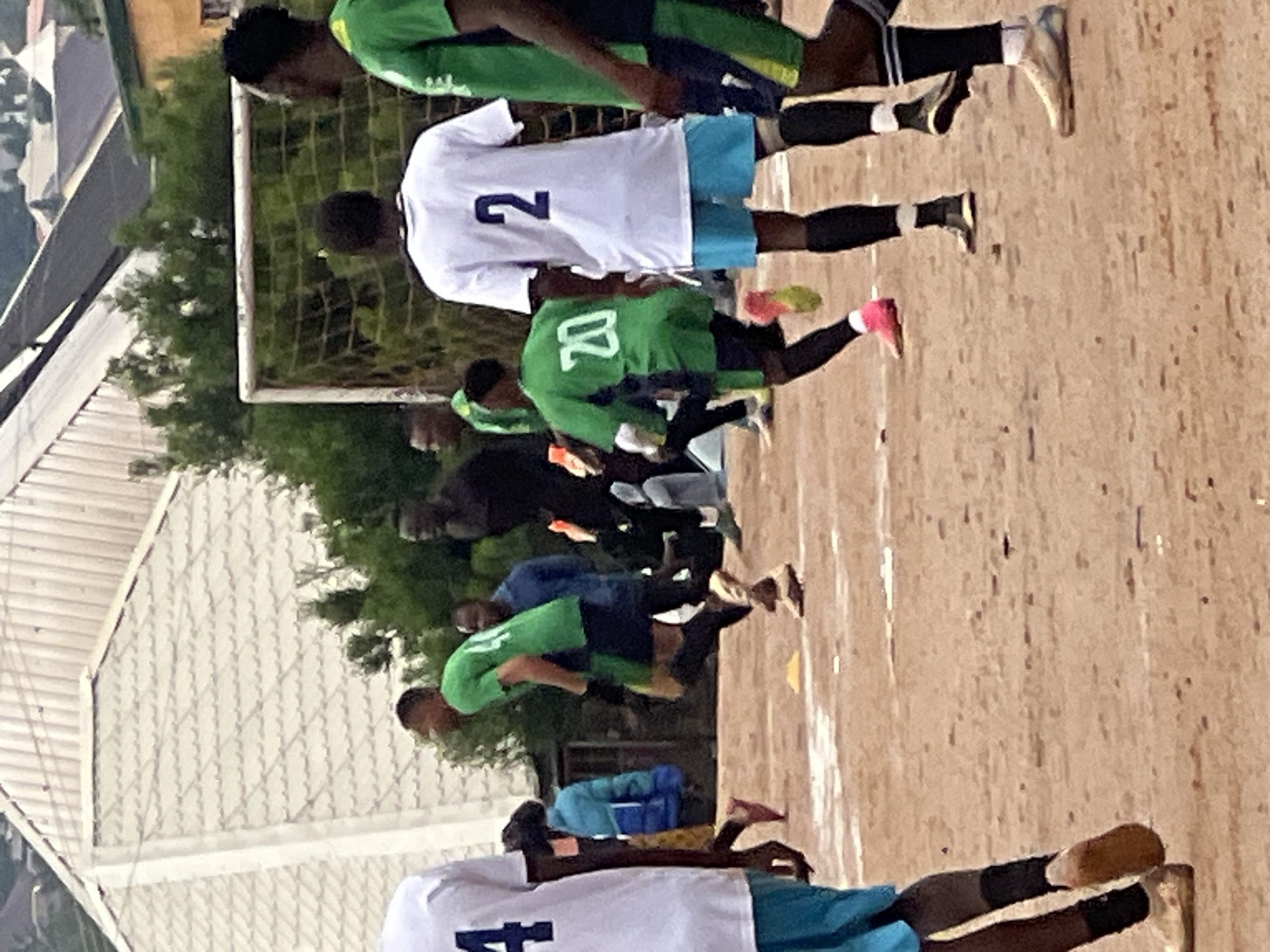 The image depicts an outdoor scene on a dirt field, likely a soccer pitch, in Jos, Nigeria. Several young men, dressed in soccer attire, are present. One individual in the foreground wears a white jersey with the number "4" on the back and blue shorts. To his right, a player in a green jersey with the number "20" is in motion, appearing to run. Further back, another player in a white jersey with the number "2" and blue shorts stands facing away from the camera. A soccer goal with netting is visible in the background, as are some trees and residential structures, including a building with a corrugated metal roof. The ground is primarily dirt with some sparse vegetation and scattered debris. The overall lighting suggests it is daytime, with overcast conditions. The presence of the soccer goal and the attire of the individuals strongly indicate a sporting event.