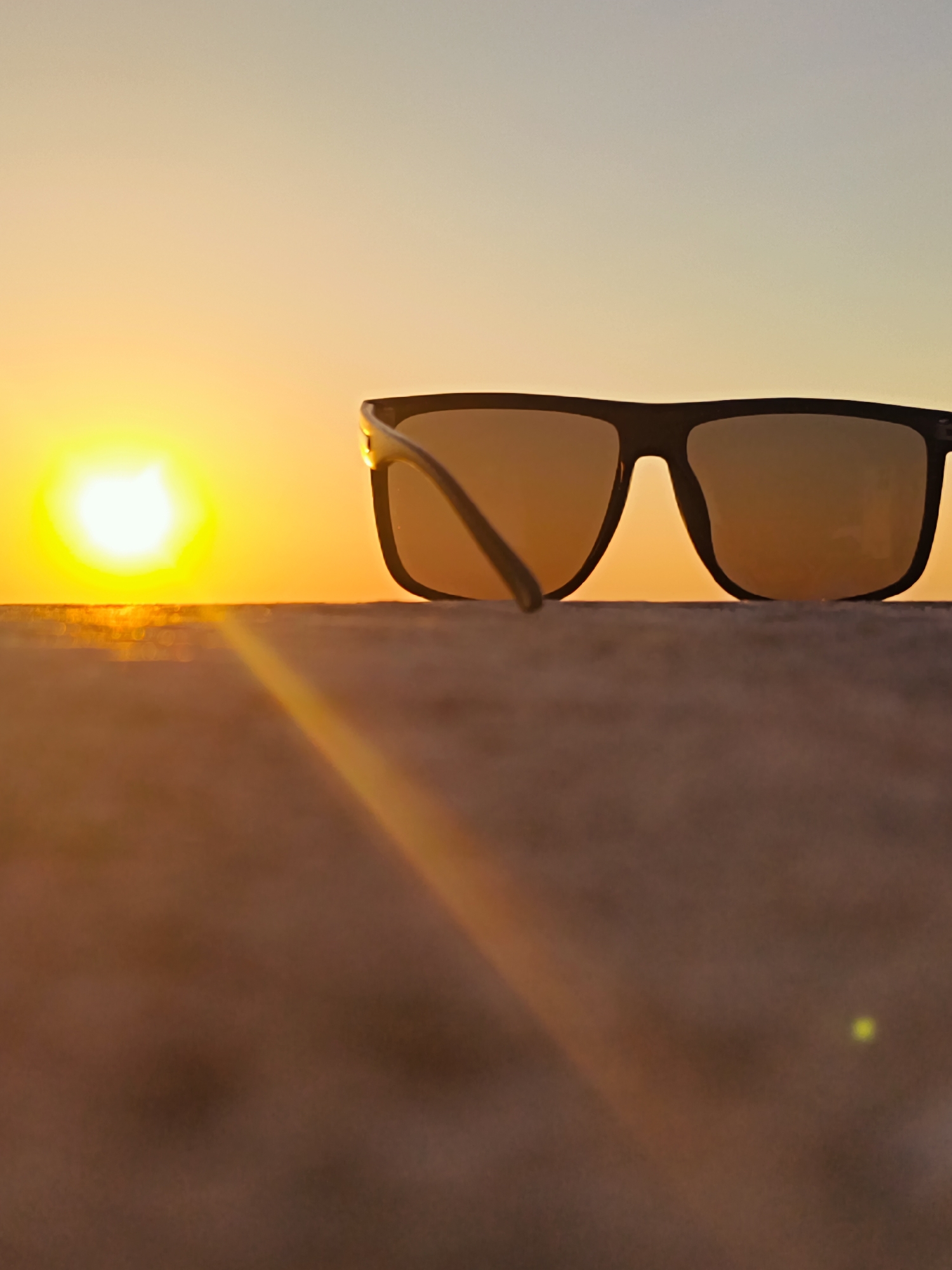 The image displays a tranquil outdoor scene dominated by a pair of sunglasses silhouetted against a radiant sunset or sunrise. The black-framed sunglasses, with lenses reflecting the warm, golden hues of the sky, are placed on a textured, flat surface that stretches across the lower half of the frame. The left arm of the sunglasses is slightly ajar.
A brilliant, luminous sun, appearing as a glowing orb, is prominently positioned on the left side of the horizon, casting an intense yellow and orange light. The sky above transitions smoothly from this vibrant warmth near the horizon to a softer, pale blue-grey at the top of the image. A distinct shimmering line is visible on the horizon to the left of the sunglasses, strongly suggesting a body of water reflecting the sunlight. Given the context of Sevastopol, Ukraine, this likely depicts a coastal vista, possibly overlooking the Black Sea.
The foreground surface is slightly out of focus, creating a sense of depth, while the sunglasses and the horizon remain sharp. A subtle lens flare is visible as a diagonal streak in the lower right portion of the image, enhancing the atmospheric effect. The overall mood is serene and warm, characteristic of a peaceful evening or early morning. There is no visible text in the image.