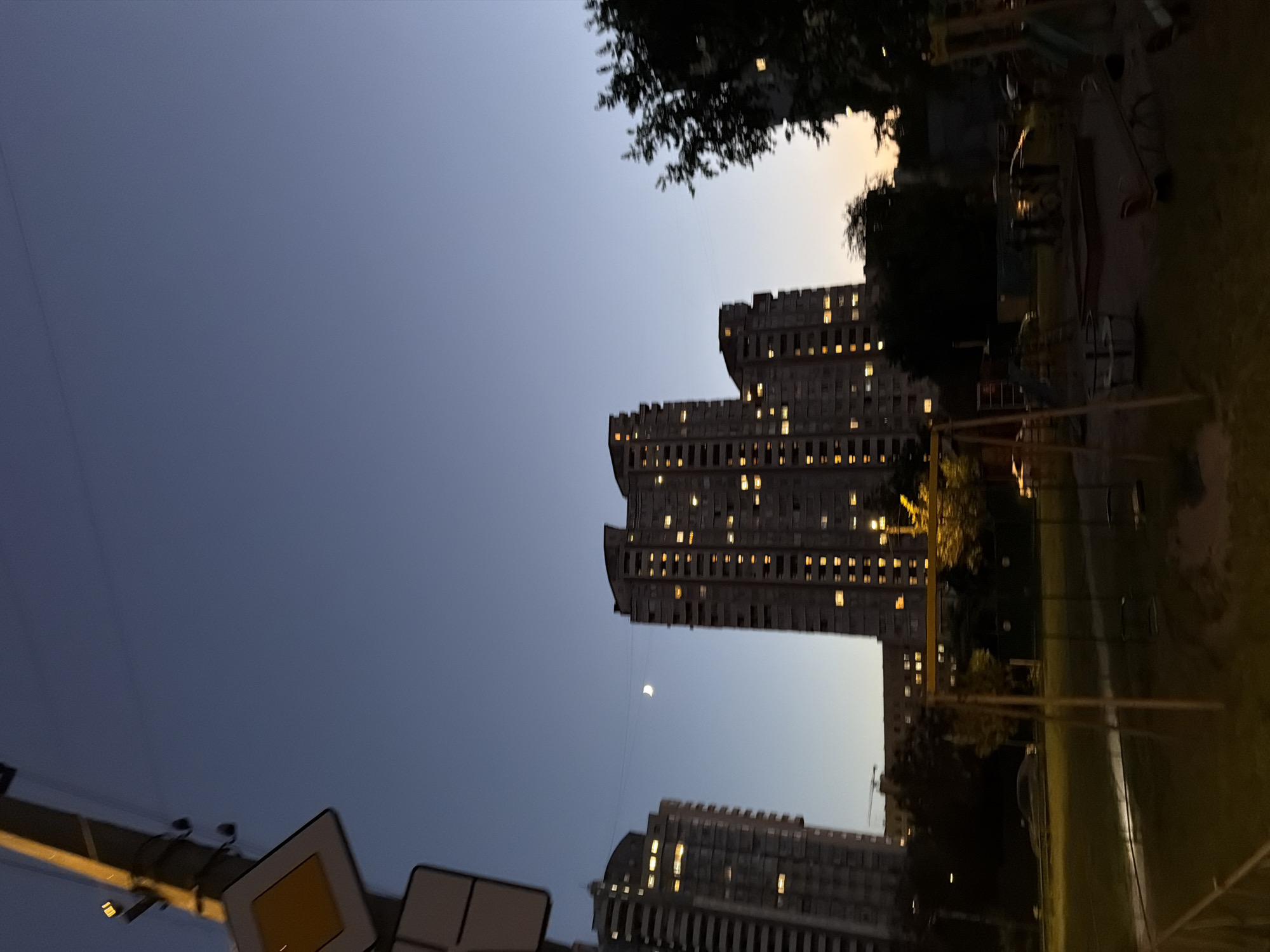 The image depicts a nighttime view of a residential area in Kyiv, Ukraine. In the foreground, a children's playground is visible with swings, a sandbox, and other play structures. A grassy area surrounds the playground. In the middle ground, several multi-story apartment buildings are lit up, indicating occupancy. The sky is dark blue, transitioning to a lighter hue near the horizon, suggesting either dusk or dawn. A crescent moon is visible in the sky between two buildings. In the far background, more buildings and trees can be observed. A street sign is present in the upper left corner of the image.