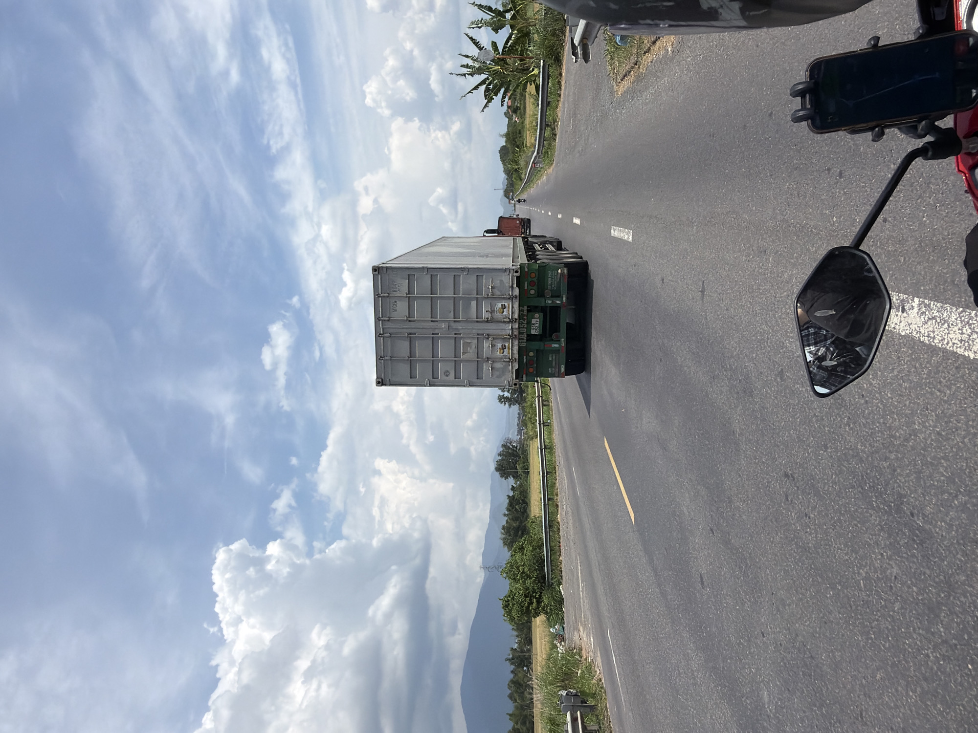 The image depicts a road scene in Dien Lac, Vietnam, under a partly cloudy sky. A large white shipping container truck is traveling on the right side of a two-lane asphalt road. The truck is followed by a red truck. In the foreground, a motorcycle's side mirror is visible, reflecting a person wearing a plaid shirt and dark pants. A smartphone is mounted on the motorcycle's handlebar. The road is lined with vegetation, including grass and trees, and a metal guardrail. In the distance, mountains are visible. The scene appears to be a bright, daytime shot, likely taken from the perspective of someone riding a motorcycle.