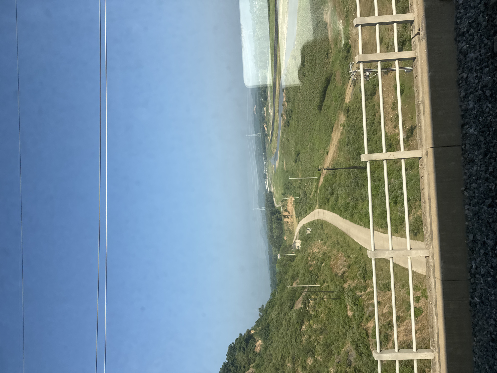The image shows a rural landscape under a clear blue sky. In the foreground, there is a concrete barrier and a white railing, suggesting the vantage point is from a bridge or elevated structure. Below the railing, a paved road winds its way up a grassy, tree-covered hill on the left.
Further into the distance, the landscape opens up to a wide, flat area with a river or body of water meandering through it. Patches of green fields and some scattered trees are visible. In the mid-ground and background, there are several utility poles and what appear to be power lines stretching across the horizon. A tall transmission tower is also visible in the distance. The overall impression is of a serene, undeveloped area with some infrastructure. It is not possible to determine the exact location within Huludao City, China, from the image alone. There are no people or animals visible in the scene.