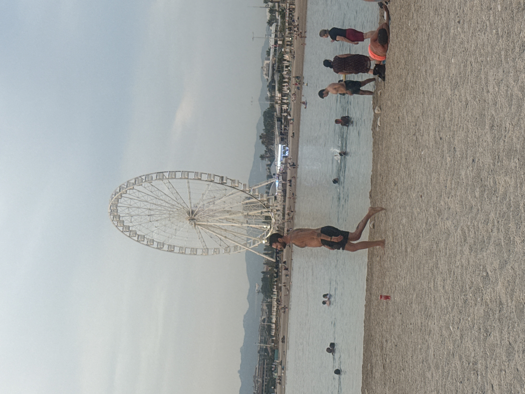 A ferris wheel stands on a beach next to the ocean. Several people are visible on the beach and in the water. Some people are wading in the ocean, while others are standing on the beach. A man in the foreground is doing a handstand on the beach. The sky is overcast, suggesting it may be a cloudy day. The scene appears to be a public beach with amenities like buildings and palm trees visible in the background.