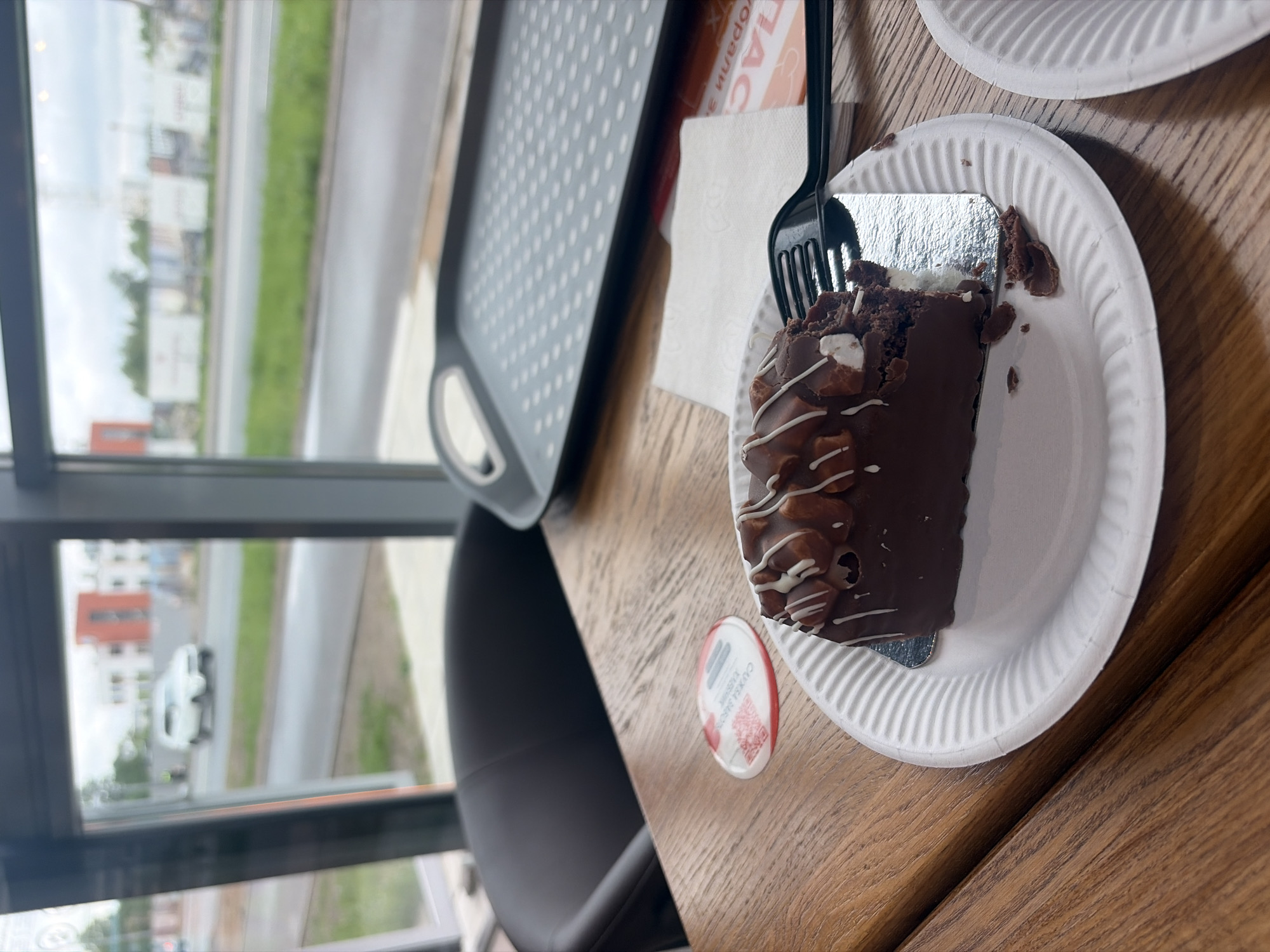 A partially eaten chocolate cake sits on a white paper plate on a wooden table next to a window overlooking a city street. A black plastic fork rests on the plate near the cake. Next to the plate is a white lid with a red stripe, likely from a yogurt or other small container. To the left of the plate is a gray tray with a hole pattern. A gray chair is partially visible in the lower left. The window shows a blurry view of buildings, trees, and a white car driving on the street outside. The location is inferred to be in Saint Petersburg, Russia, based on the text on the lid which looks like the Cyrillic alphabet.