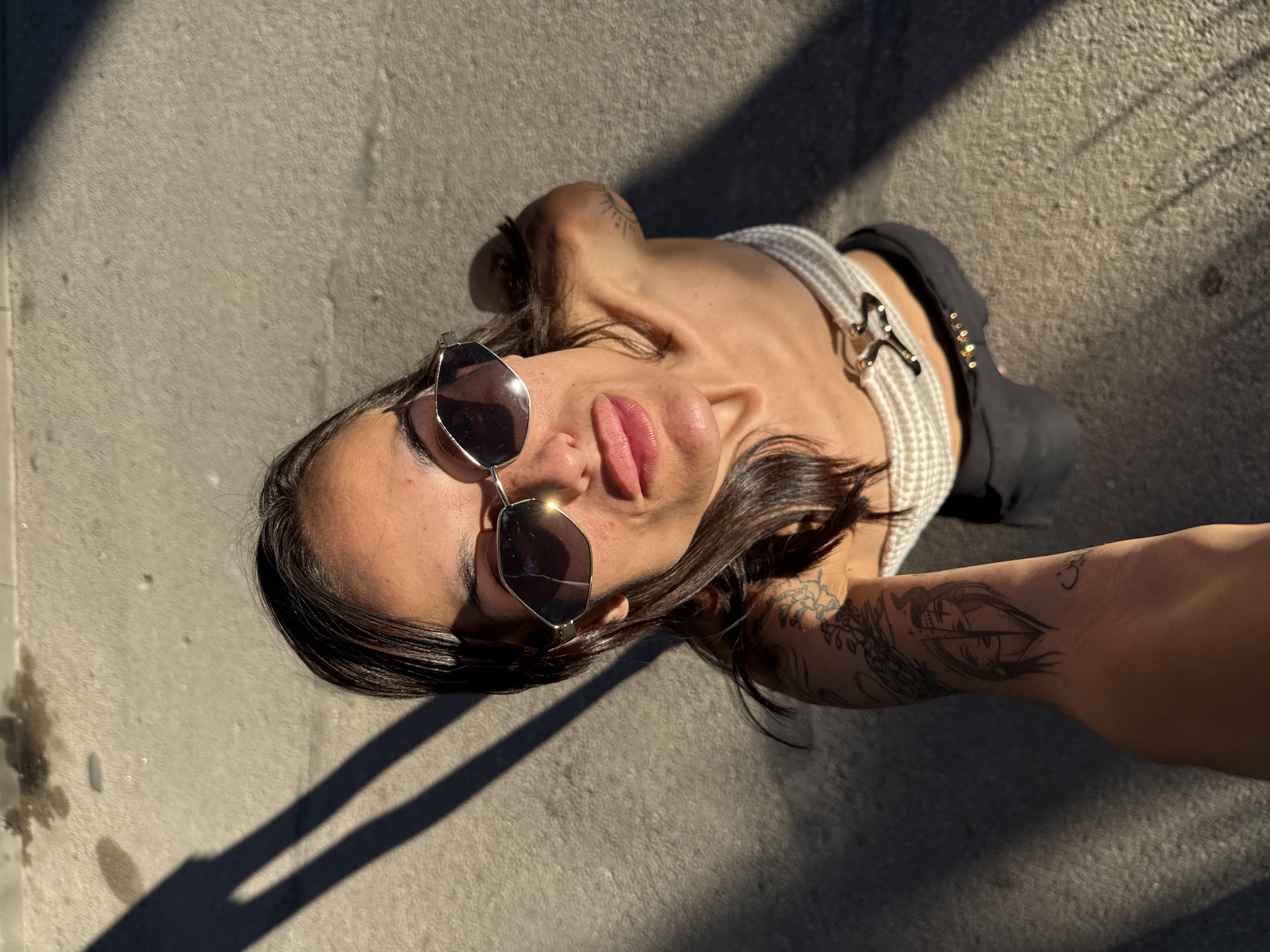 A woman is captured in a high-angle, selfie-style shot outdoors. She is wearing sunglasses, a strapless top, and dark pants. Her left arm is extended, displaying tattoos. The ground beneath her appears to be pavement or concrete, with a shadow cast diagonally across the scene, indicating it is likely daytime. The image does not contain any information that can be used to determine the location within the city of Pleven, Bulgaria.