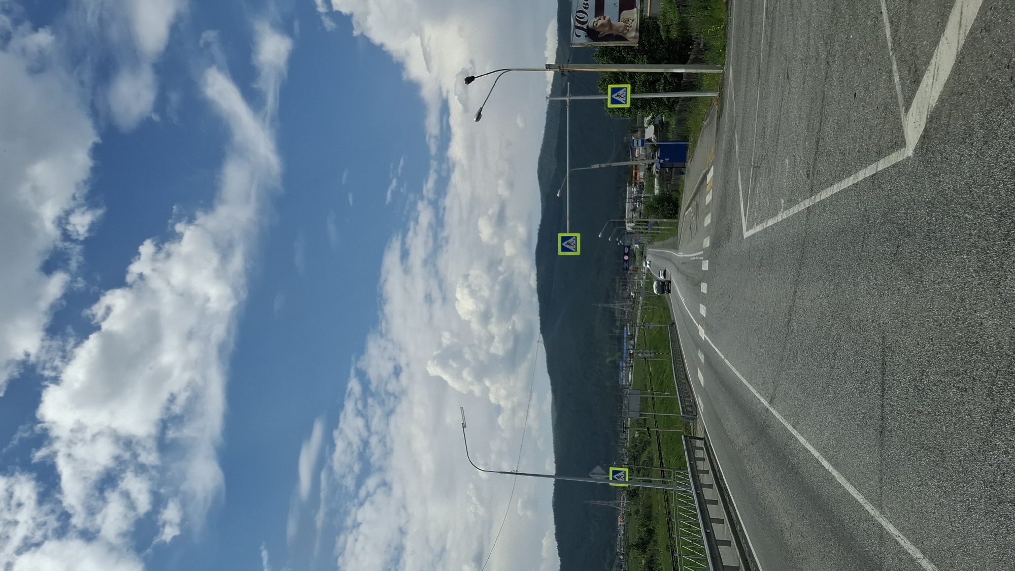 This is a vertically oriented, eye-level, outdoor shot looking down a paved road in Kultuk, Russia, on a partly cloudy day. The road is asphalt, with white lane markings and a pedestrian crossing marked with white stripes. On the left side of the road, there's a metal guardrail. In the distance, there are mountains covered in trees, and on the right side of the road, a billboard with a woman's face on it. There are streetlights and pedestrian crossing signs along the road. Two vehicles, a truck and a car, are visible driving on the road in the distance. The sky is blue with white, fluffy clouds.