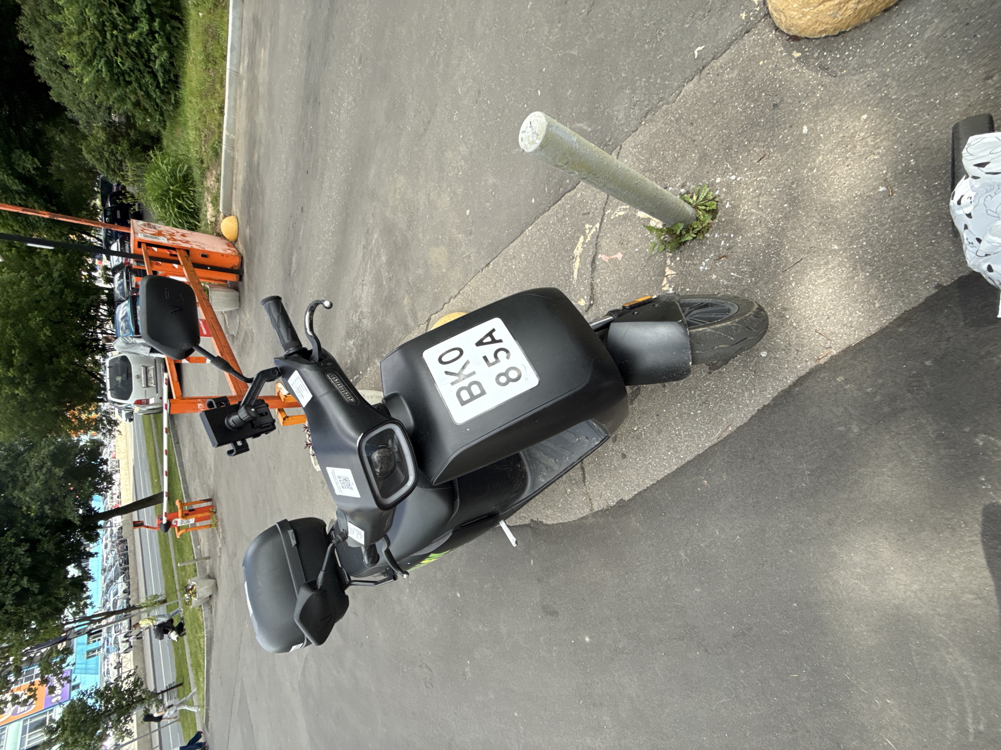 The image shows an electric scooter parked on a paved surface next to a metal pole. The scooter is black and has a license plate that reads "BK0 85A". The scene appears to be outdoors, with a grassy area and trees visible in the background. The lighting suggests it is daytime. The scooter is the primary object of focus, and no people are clearly visible. The setting appears to be urban or suburban, possibly a parking lot or sidewalk. The image is oriented vertically. There are some orange barriers and a parking gate visible in the background, suggesting a controlled access area.