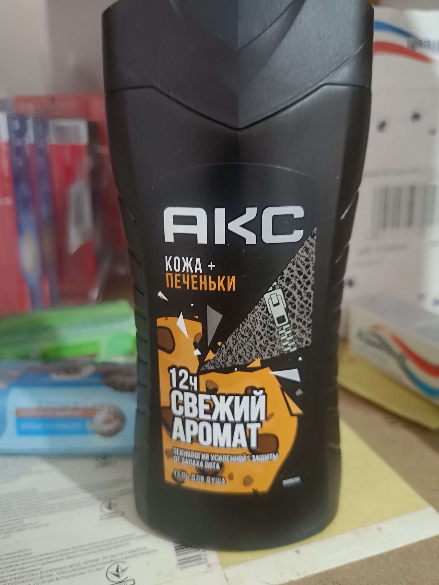 A black plastic bottle with a curved top is positioned slightly off-center in the foreground of the image, angled towards the left. The bottle has a textured surface and features text and graphics in white, orange, and grey. Prominently displayed on the bottle is the word "АКС" in large white letters. Below this, in smaller white and orange text, are the words "КОЖА + ПЕЧЕНЬКИ". A graphic of a zipper is visible on the right side of the bottle, overlaid on a textured grey area. Further down, a large orange graphic resembling cheese with holes contains the text "12Ч СВЕЖИЙ АРОМАТ". Below this, in smaller white text, is "ТЕХНОЛОГИЯ УСИЛЕННОЙ ЗАЩИТЫ ОТ ЗАПАХА ПОТА" and "ГЕЛЬ ДЛЯ ДУША". A small alphanumeric code, "93030604", is also present. The background is blurred, but appears to be indoors with shelves and various objects, suggesting a retail or storage environment. There is no indication of the city of Berdiansk, Ukraine, within the image.